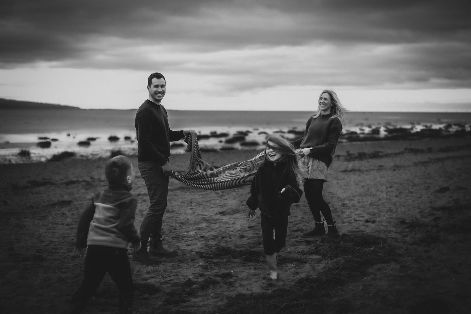 Black and white family photo on a rocky beach, parents holding a blanket with two young children playing outdoors.