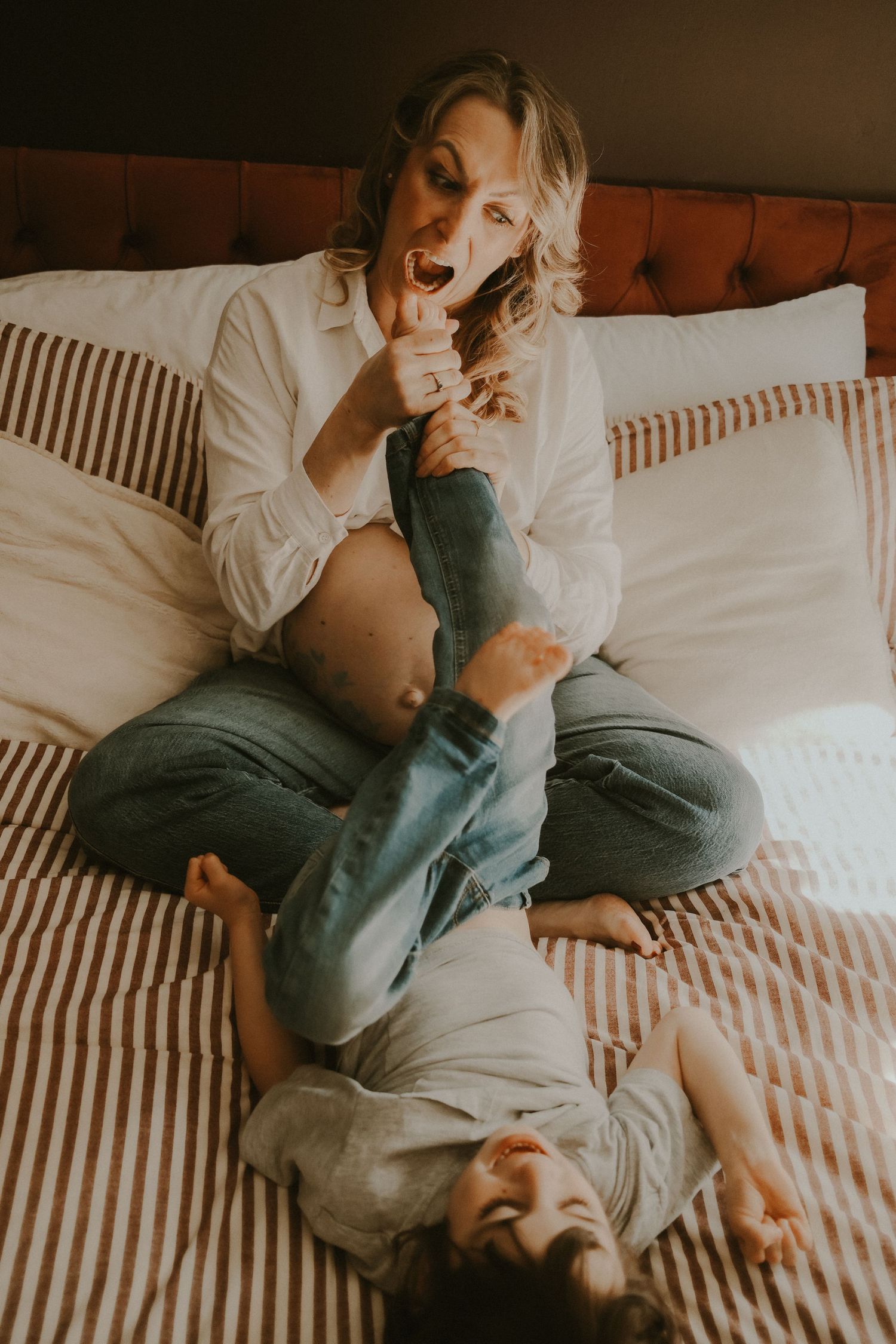 Pregnant woman sitting cross-legged on striped bed, playfully holding toddler's legs up while laughing together.