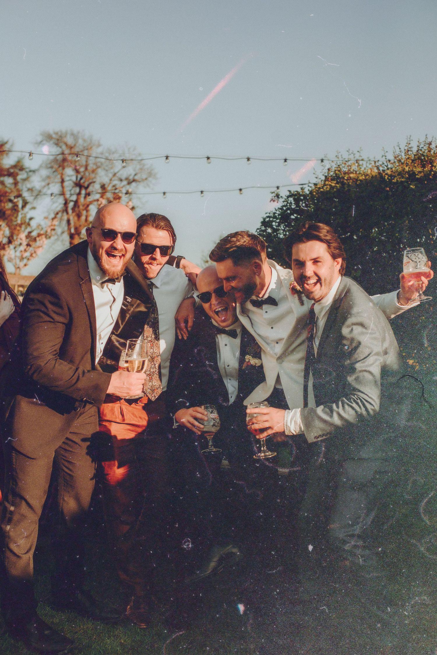 Four men in formal suits celebrating outdoors at a wedding, laughing and holding champagne glasses.