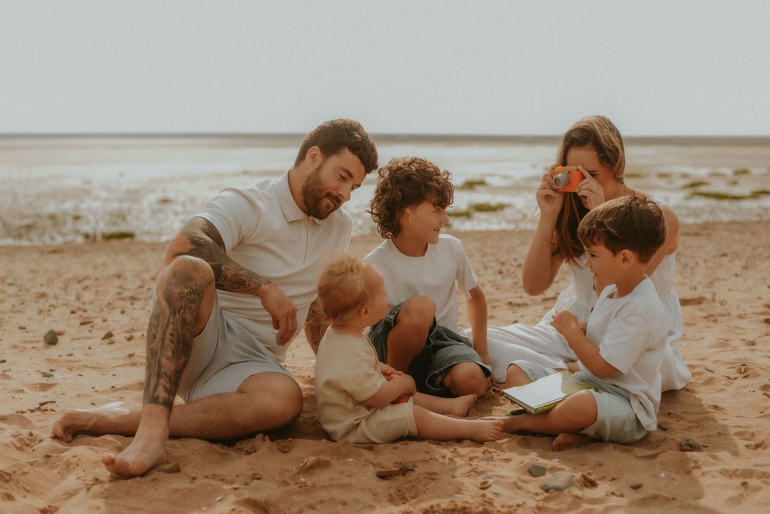 Family of five enjoying a sunny beach day, sitting together on golden sand with ocean waves in the background.