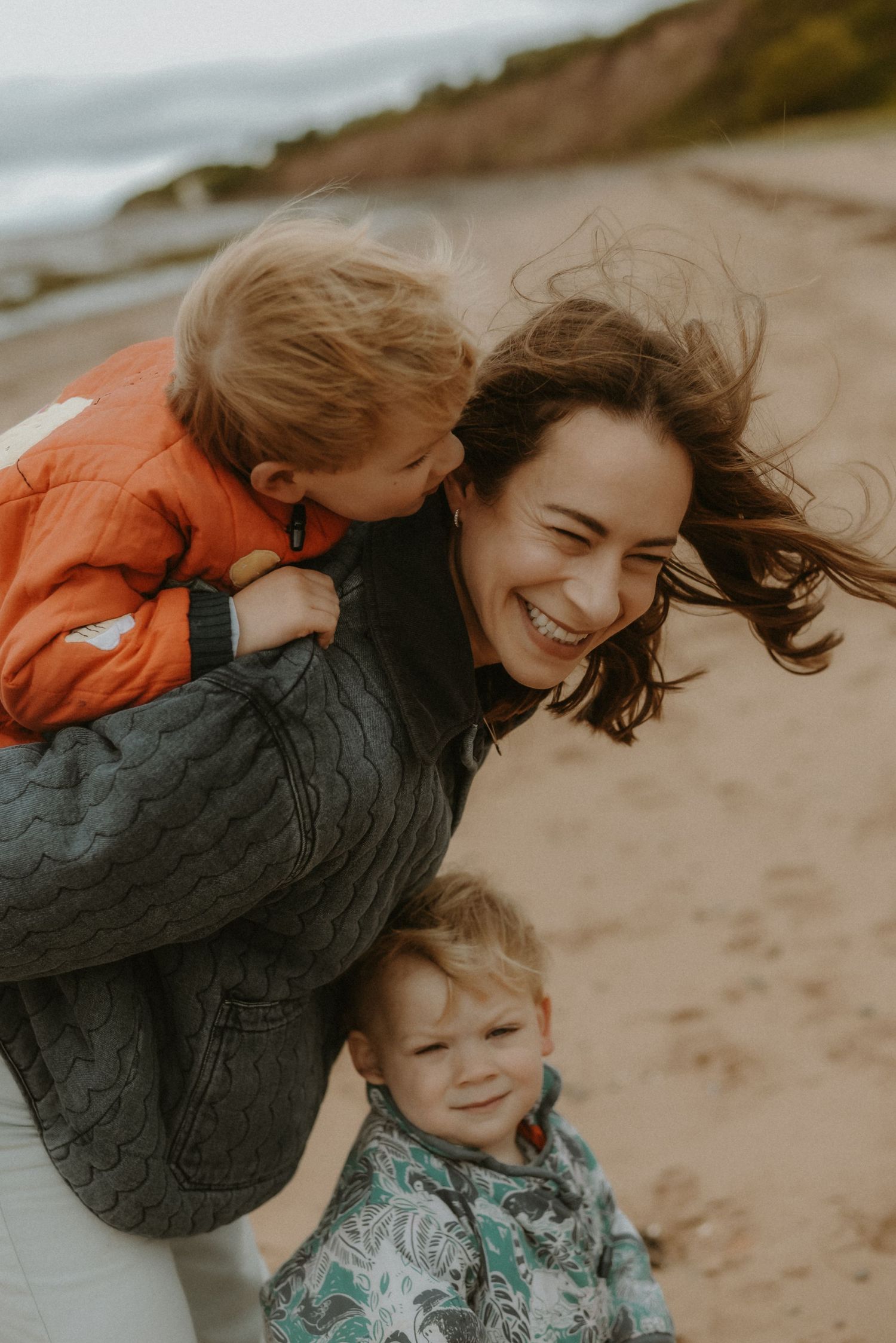 A laughing woman carries a toddler in an orange jacket on her back at a beach, while another young child smiles nearby.