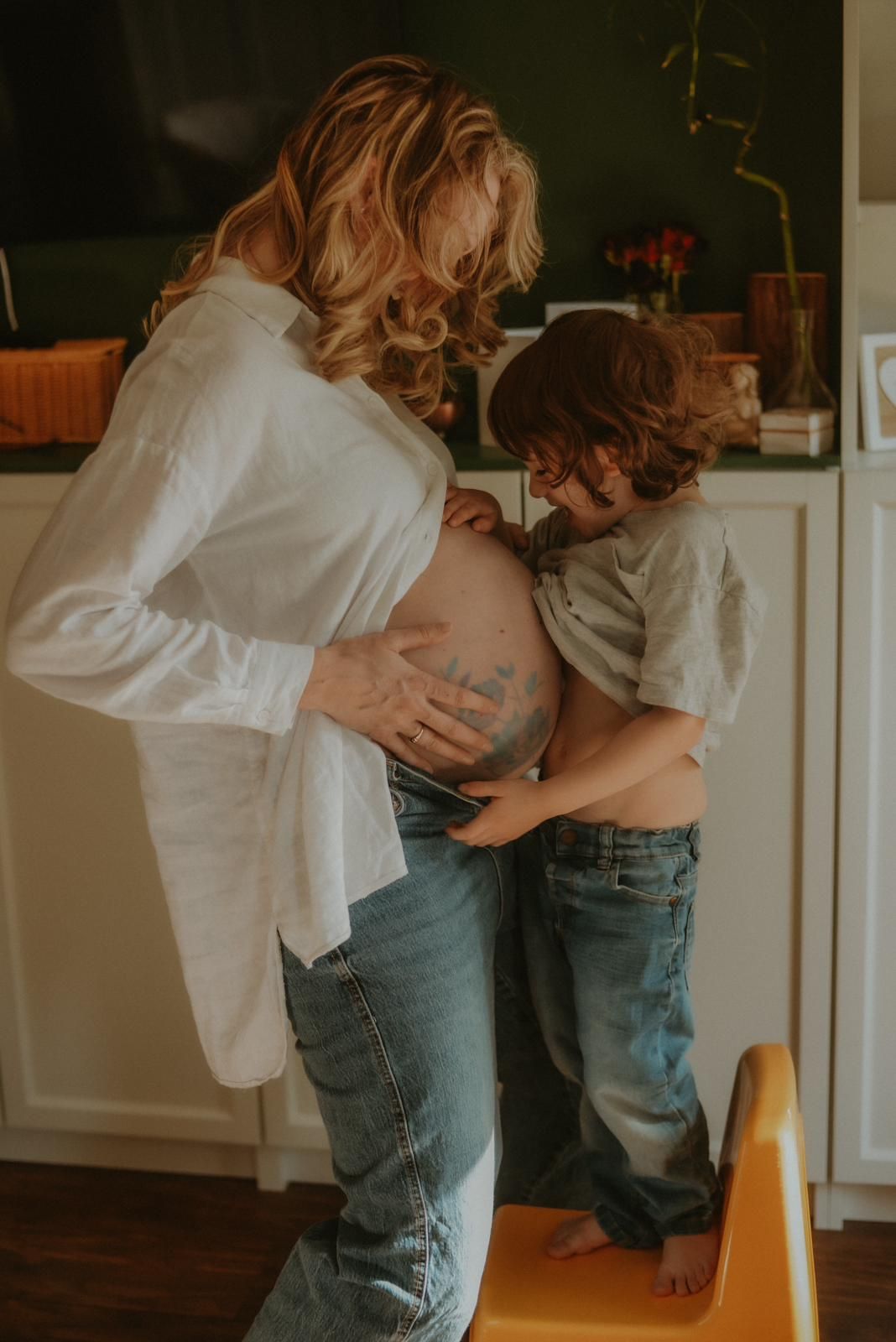 Pregnant mother lifting shirt as young child stands on stool to listen to her baby bump.