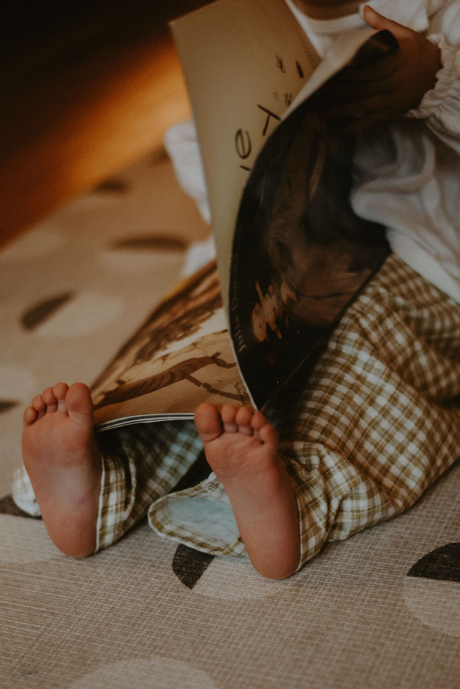 Close-up of a baby's tiny bare feet peeking out from plaid pants while reading a book.