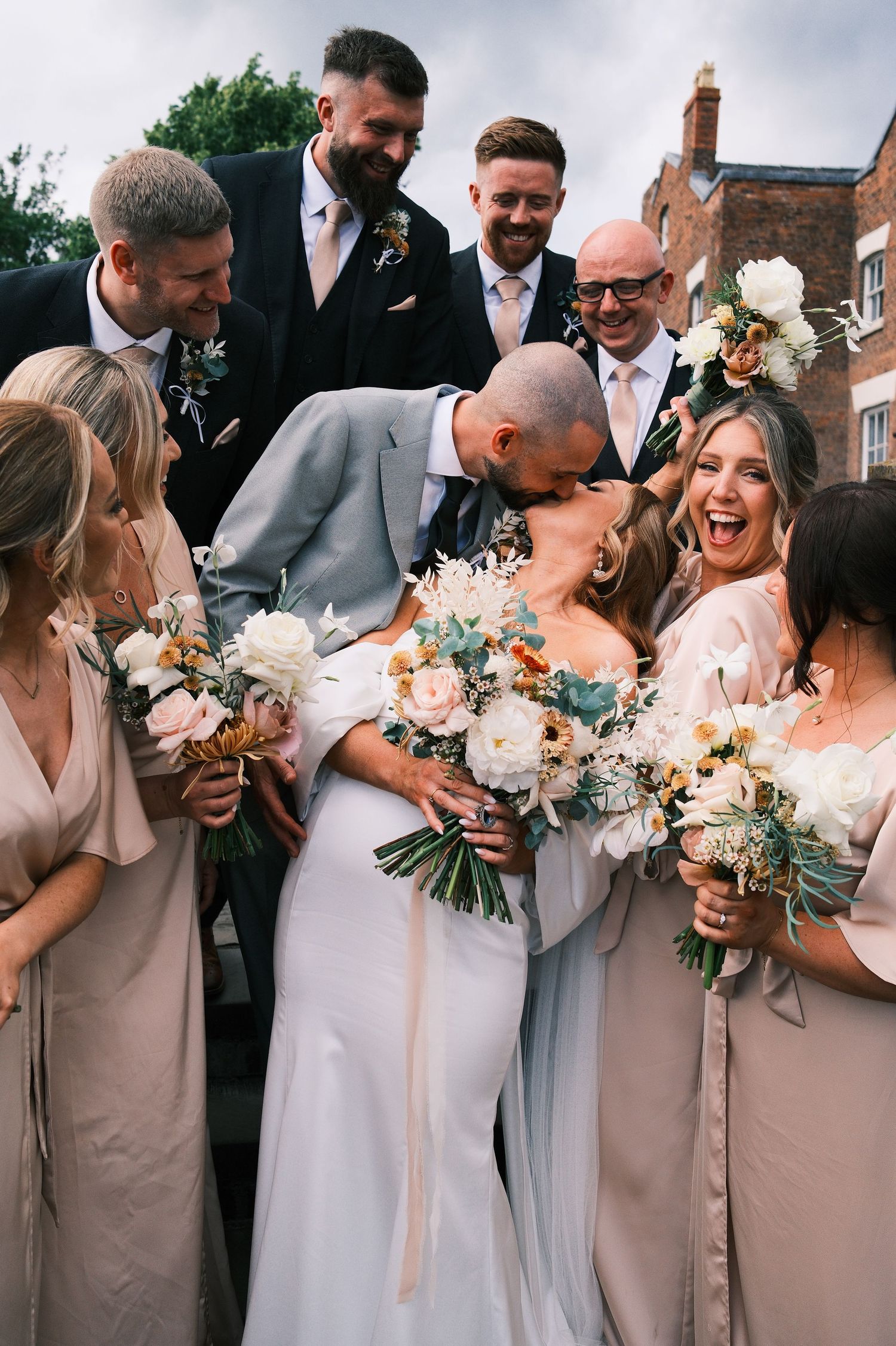 Bride and groom share a kiss surrounded by smiling bridesmaids in blush gowns and groomsmen holding bouquets at a wedding.