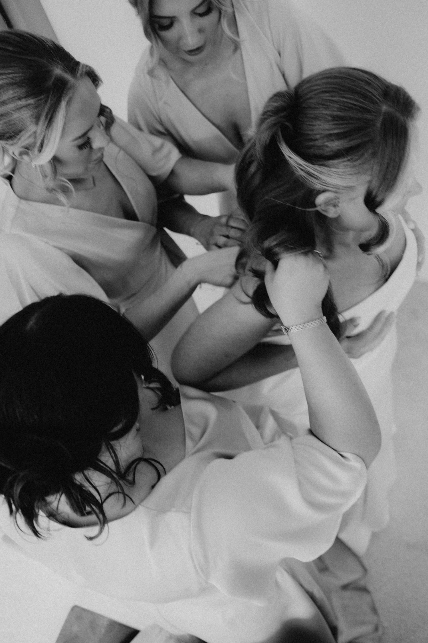 Bridesmaids in white robes helping bride with hair in black and white photo.