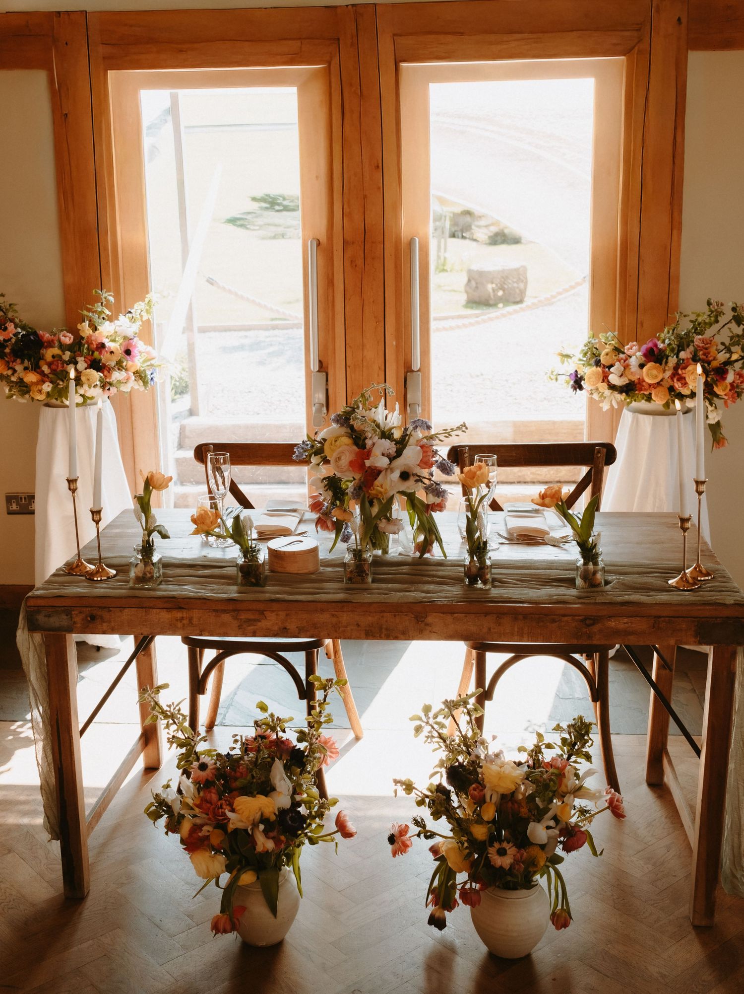 Rustic wooden sweetheart table adorned with colorful floral arrangements in warm tones, set before large wooden-framed windows.