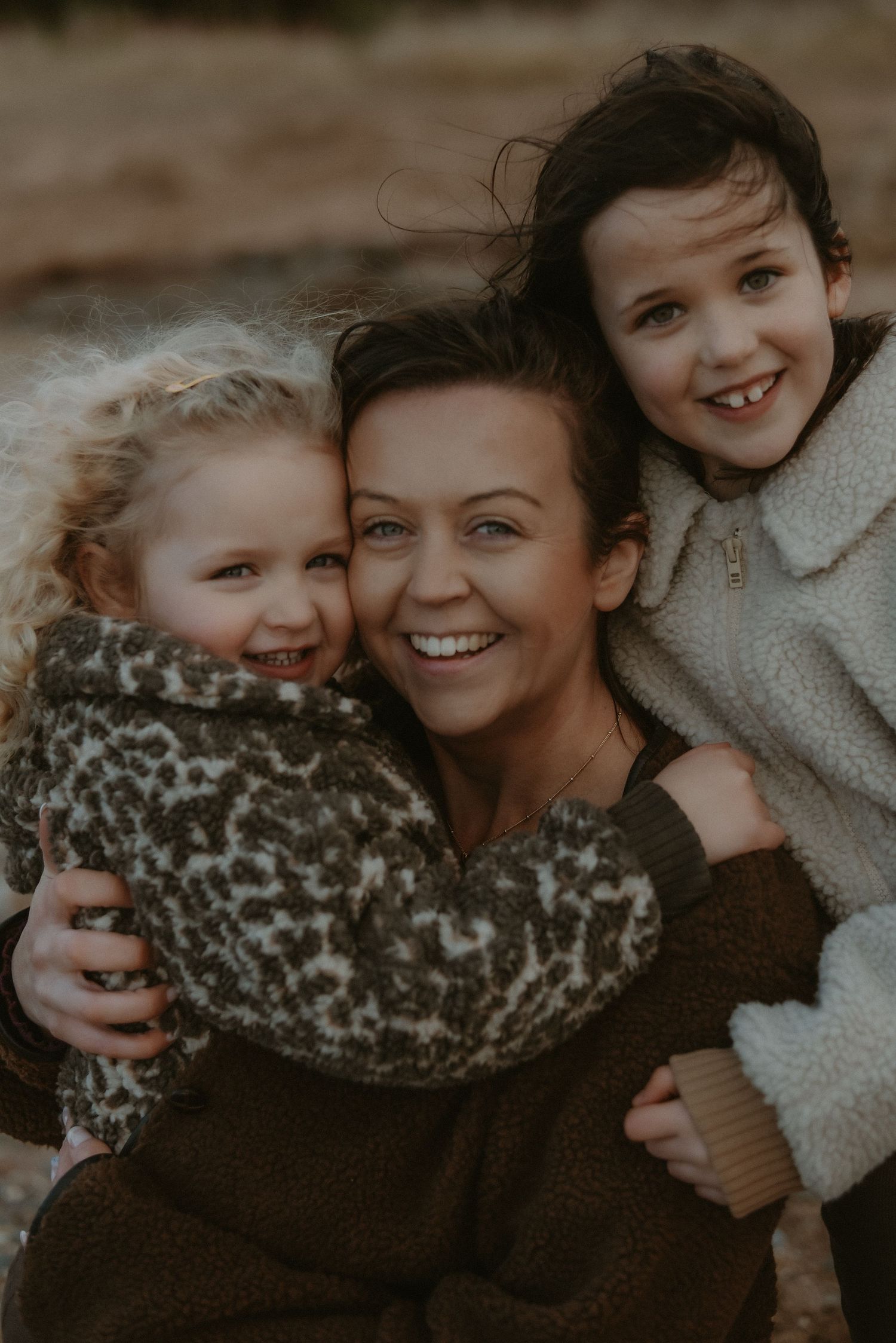 Smiling mother embraced by two young daughters outdoors in warm autumn clothing.