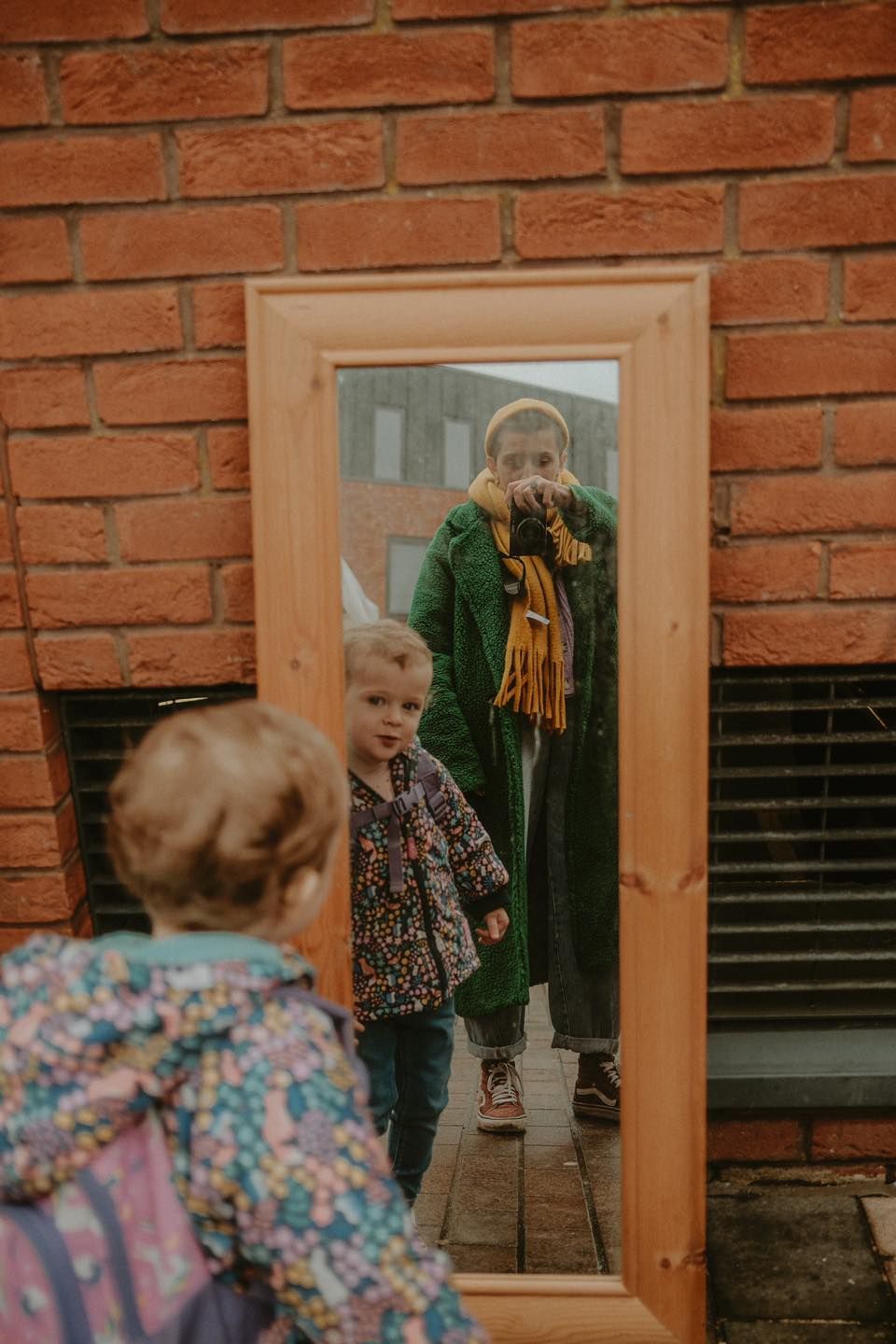 A child and adult reflected in a wooden-framed mirror against a red brick wall.
