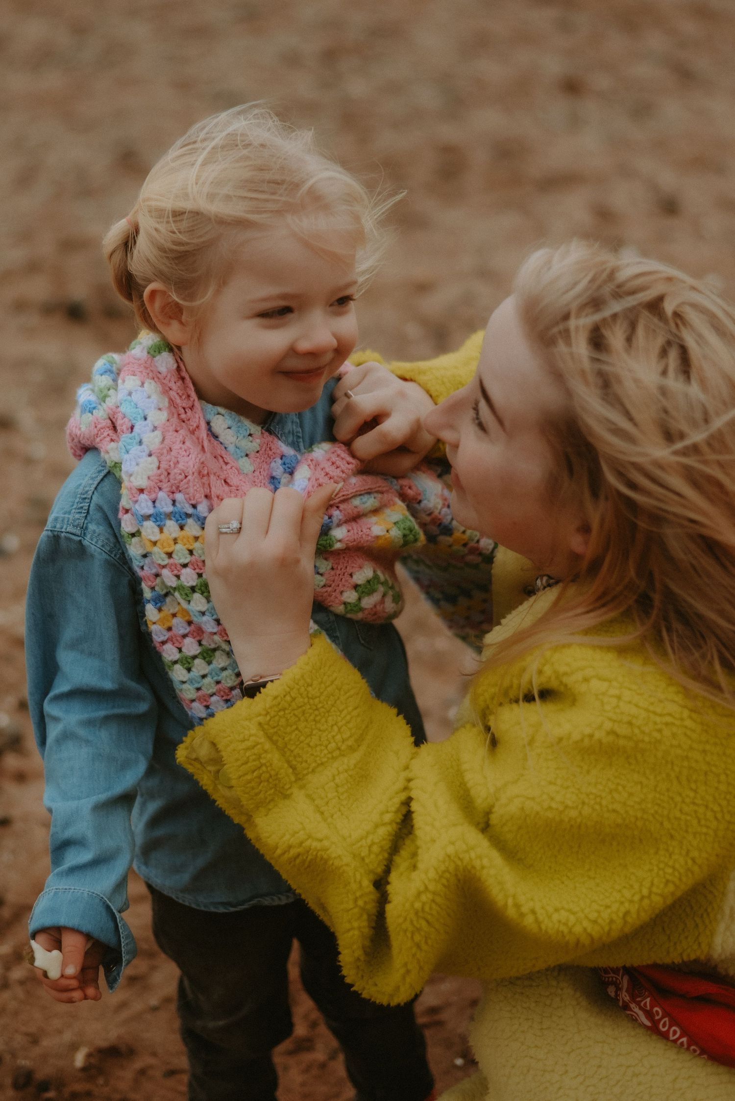 A smiling mother in a yellow jacket adjusting her daughter's colorful crochet scarf outdoors.