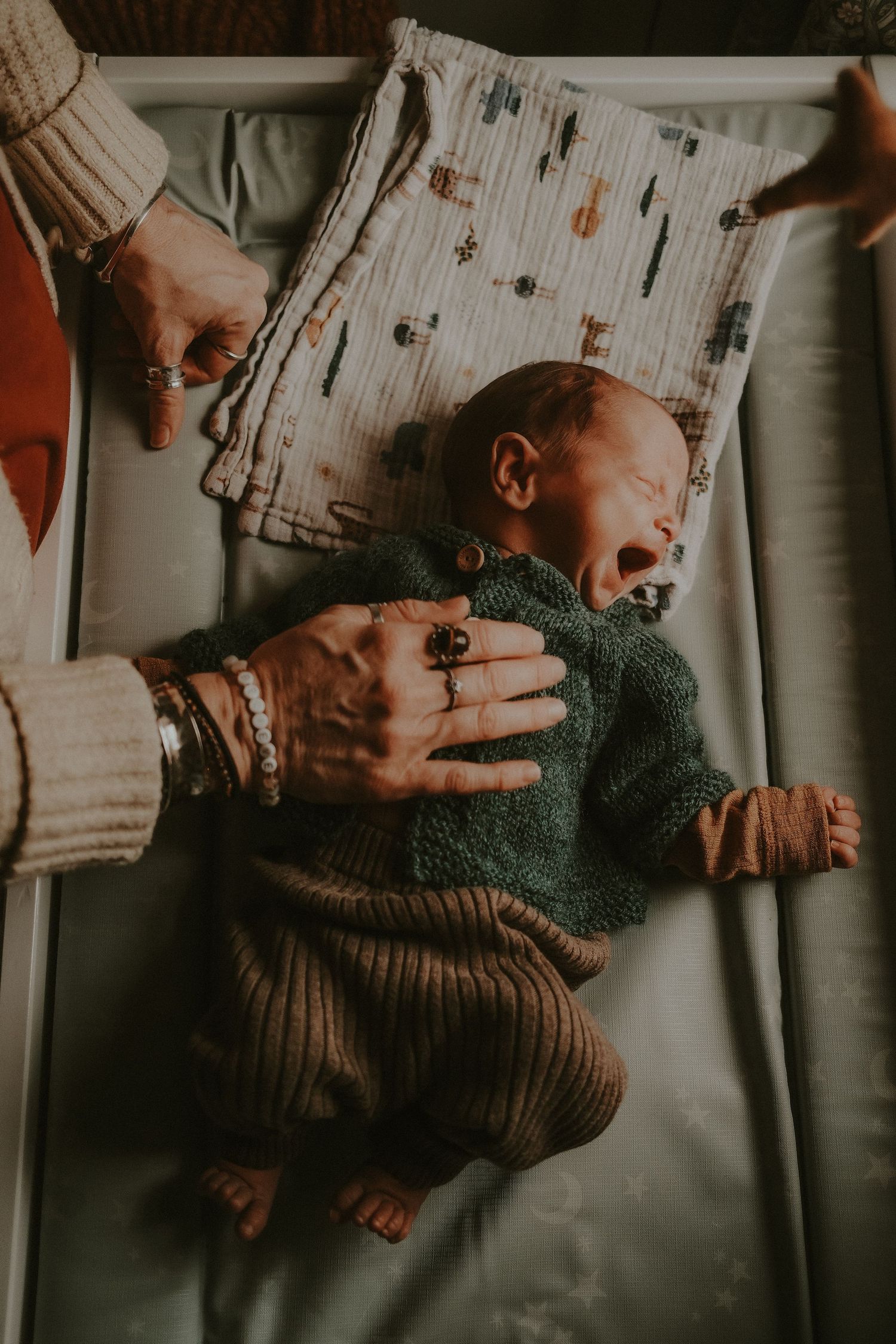 Newborn baby in green knit sweater lying in crib while adult hands with rings gently rest on chest.