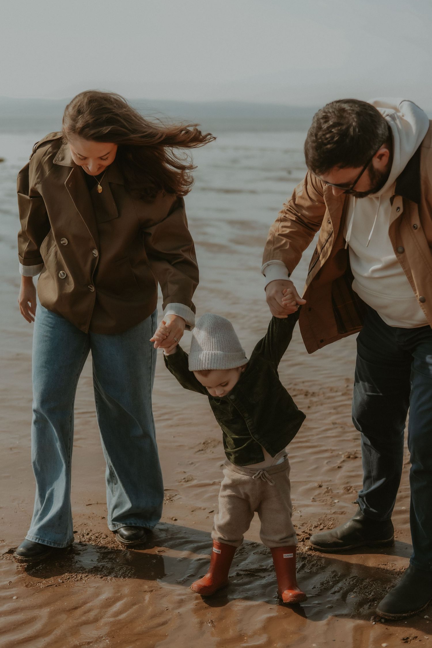 Parents holding hands with toddler in red boots walking on wet sandy beach.