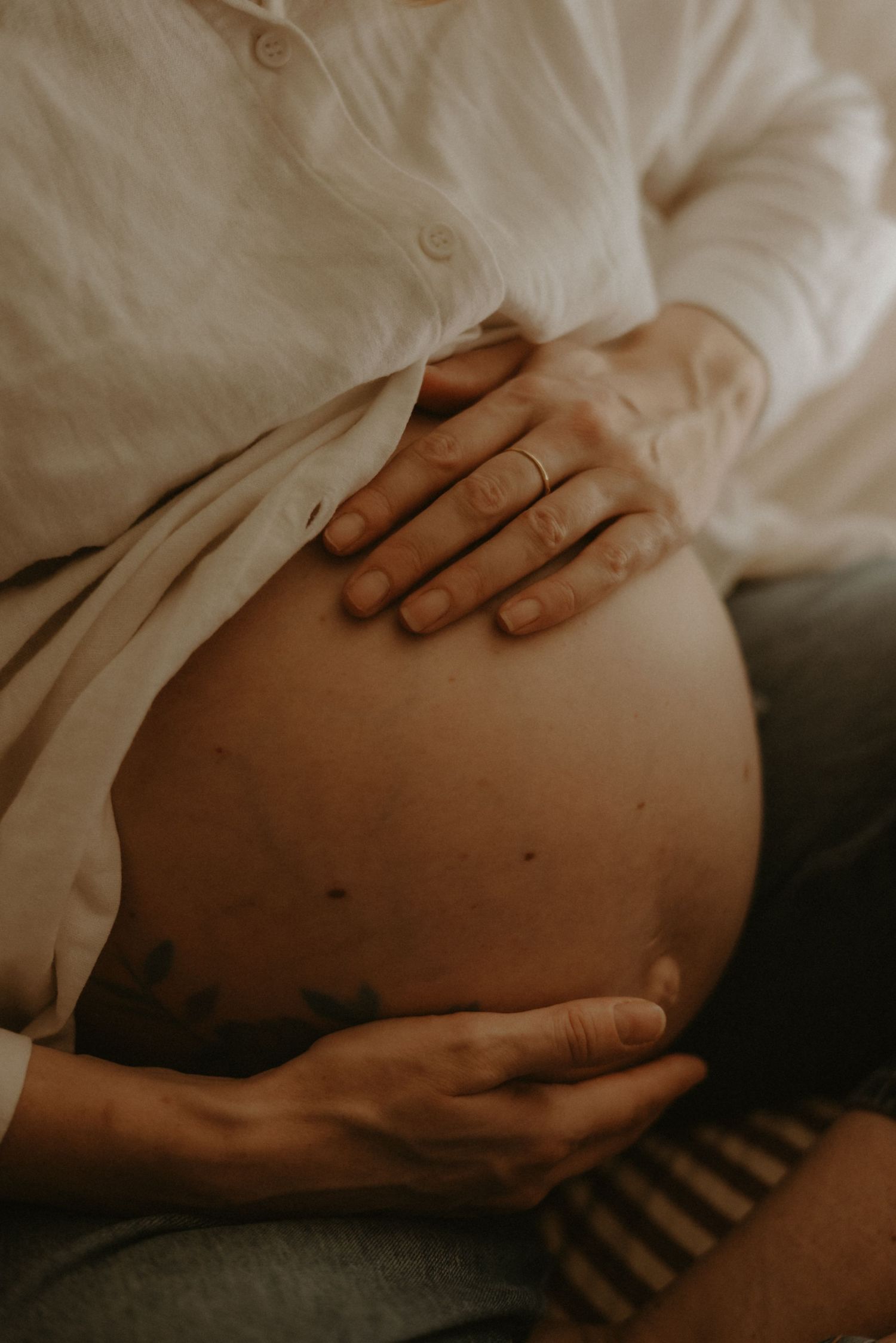 Close-up of pregnant woman in white shirt cradling bare baby bump with both hands.