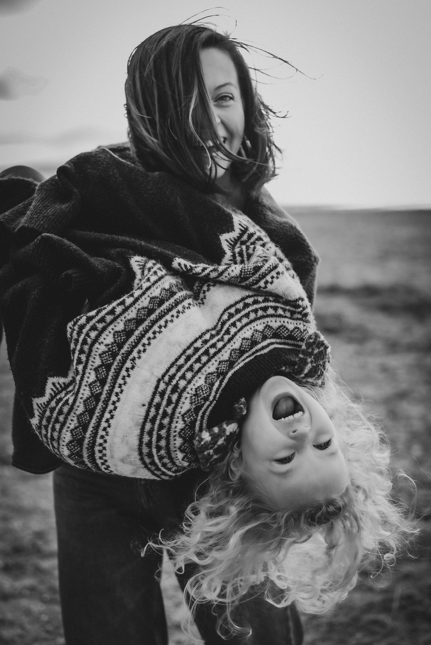 Black and white image of smiling woman playfully holding child upside down outdoors.