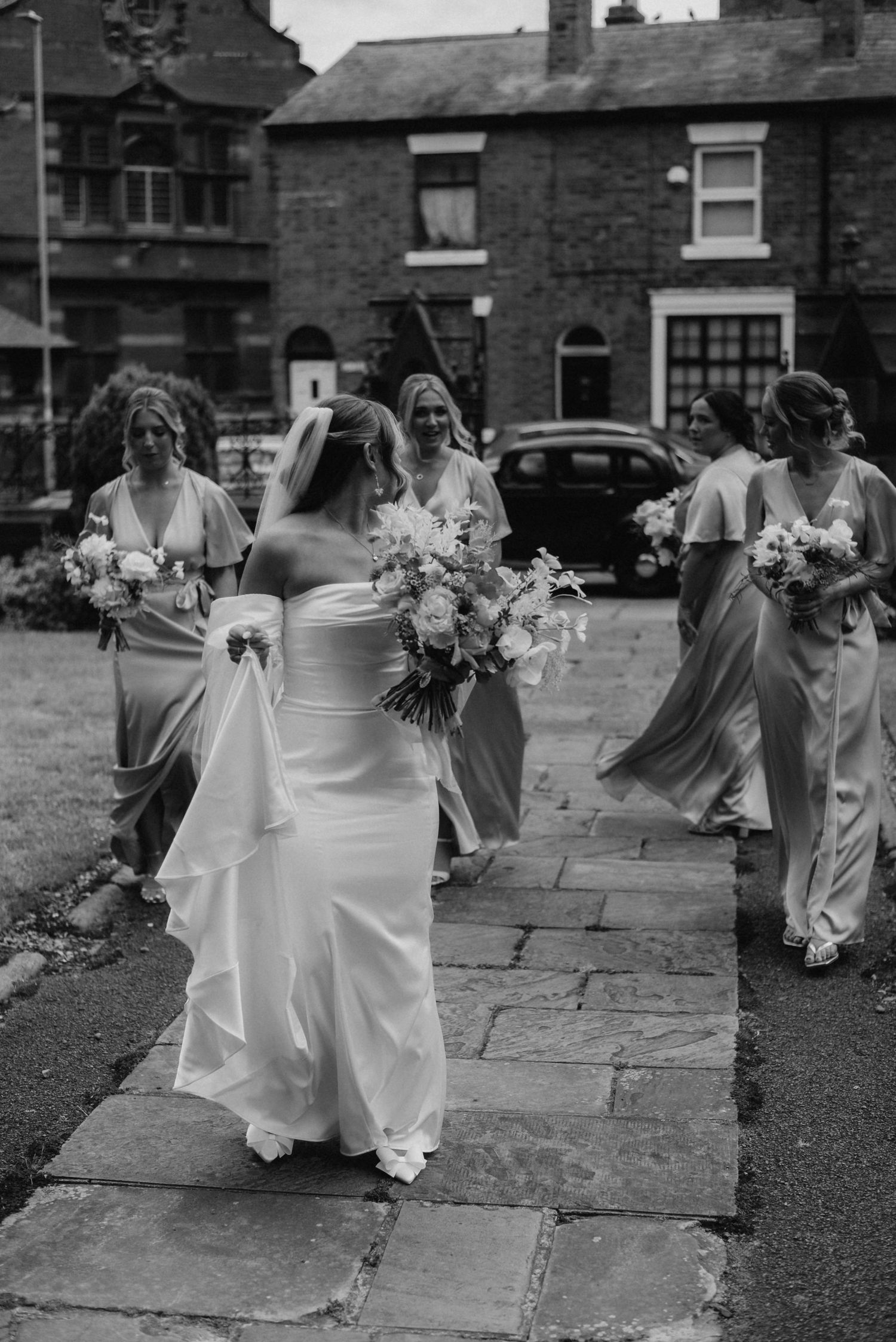 Bride in white gown walks with bridesmaids in flowing dresses, holding bouquets on stone path, black and white wedding photo.