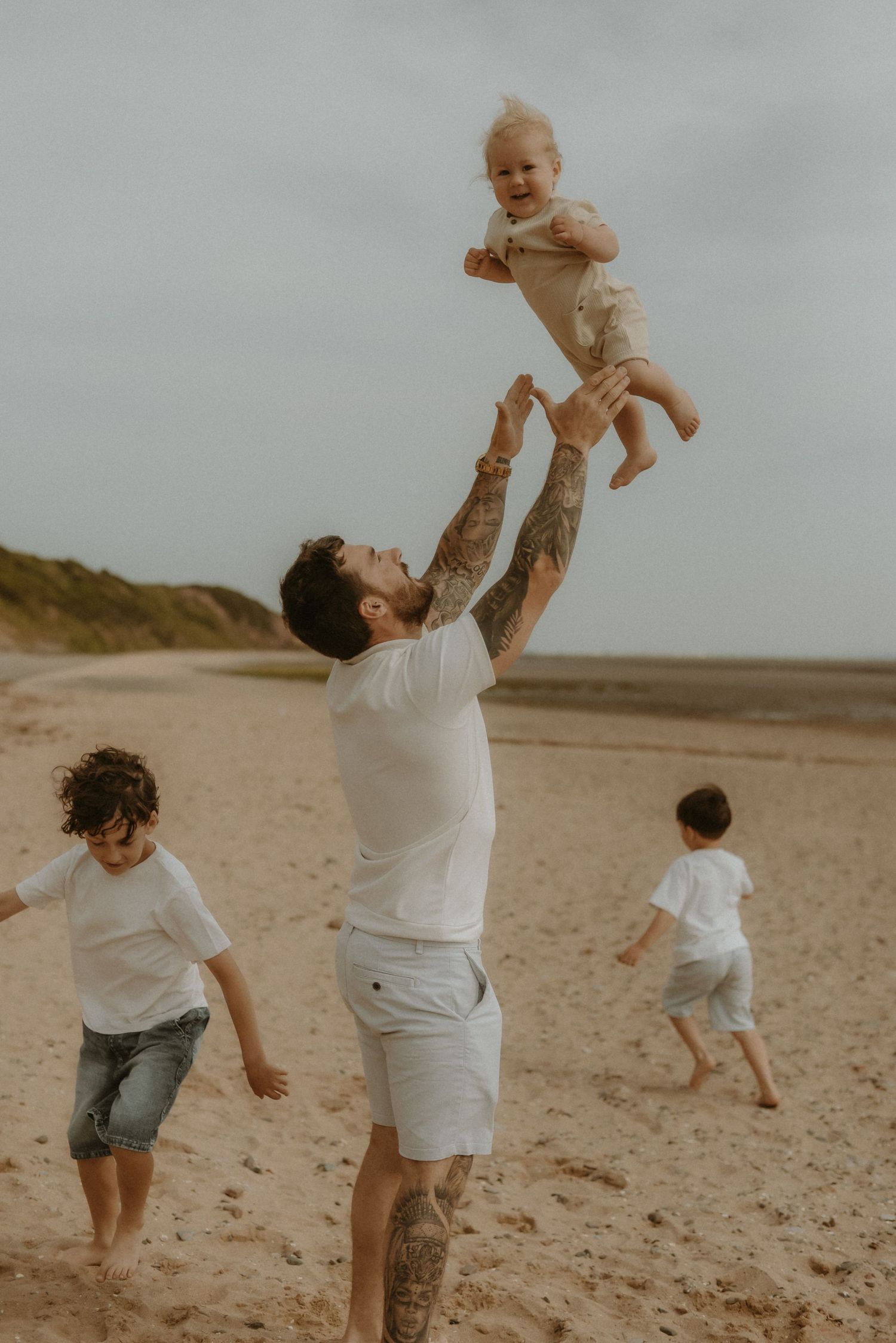 Father tossing baby in air on sandy beach while two young children play nearby, all wearing white outfits.