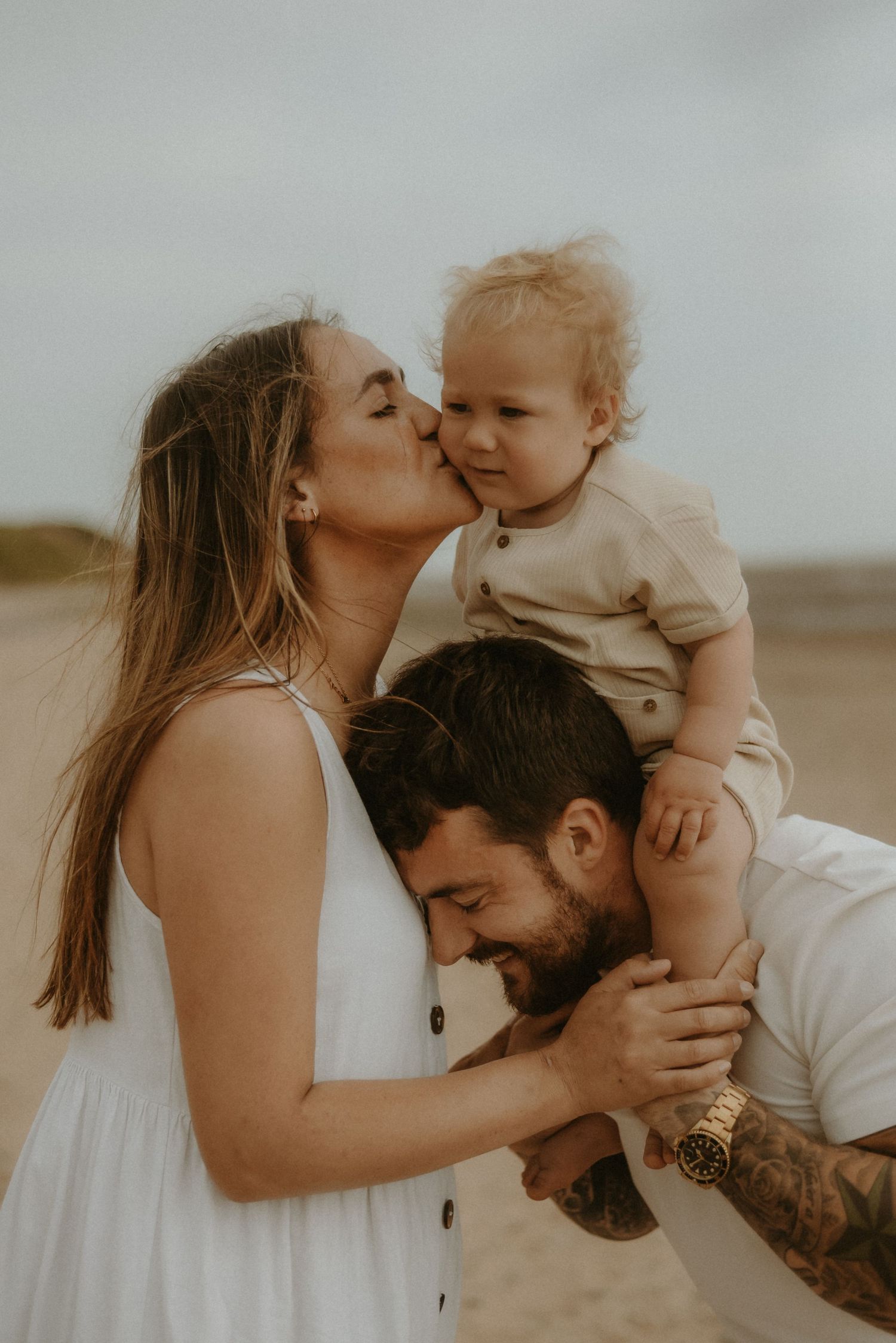 A young family shares a tender moment on the beach, with mom kissing their toddler perched on dads shoulders.