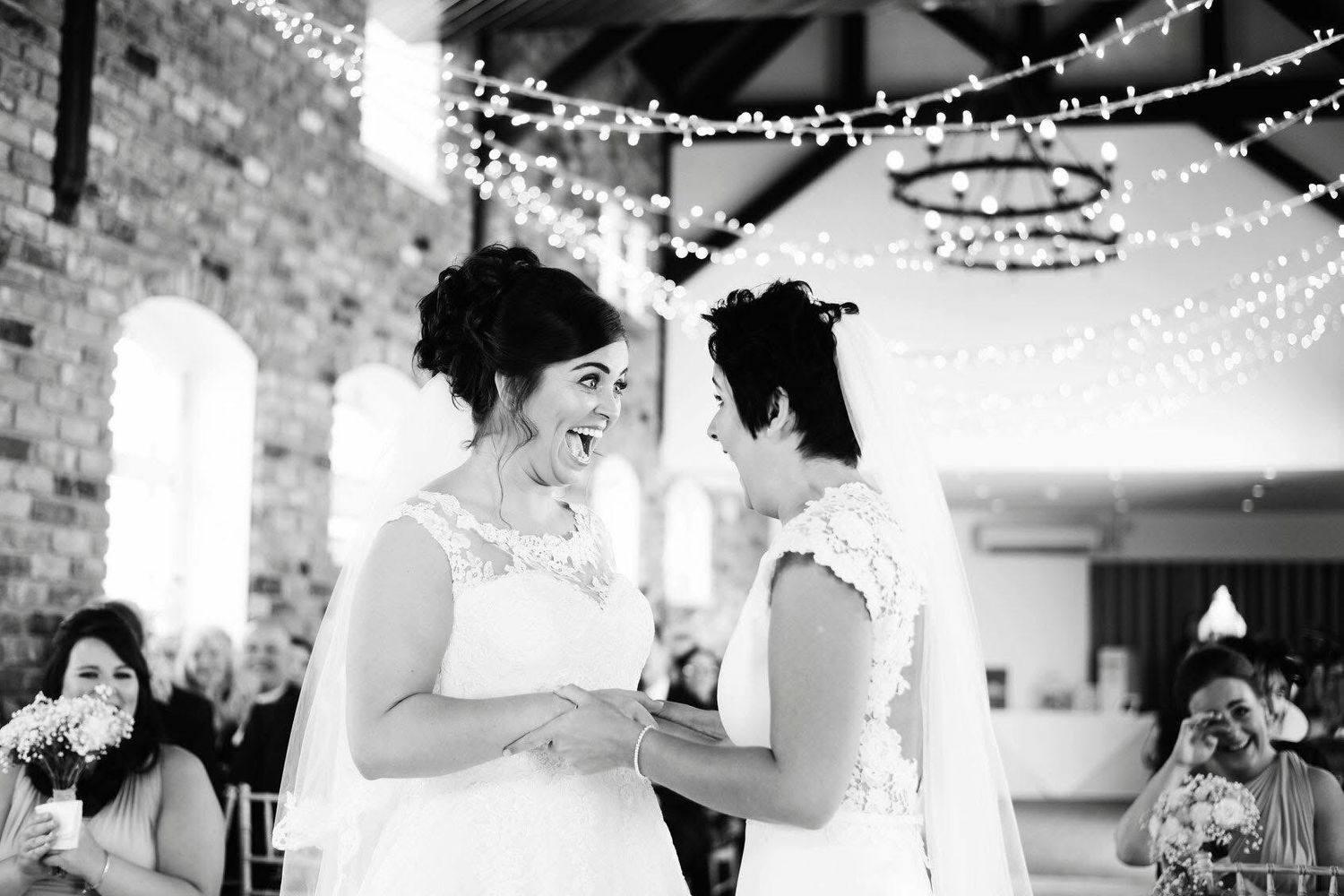 Two brides share a joyful moment during their wedding ceremony in a rustic venue decorated with fairy lights.