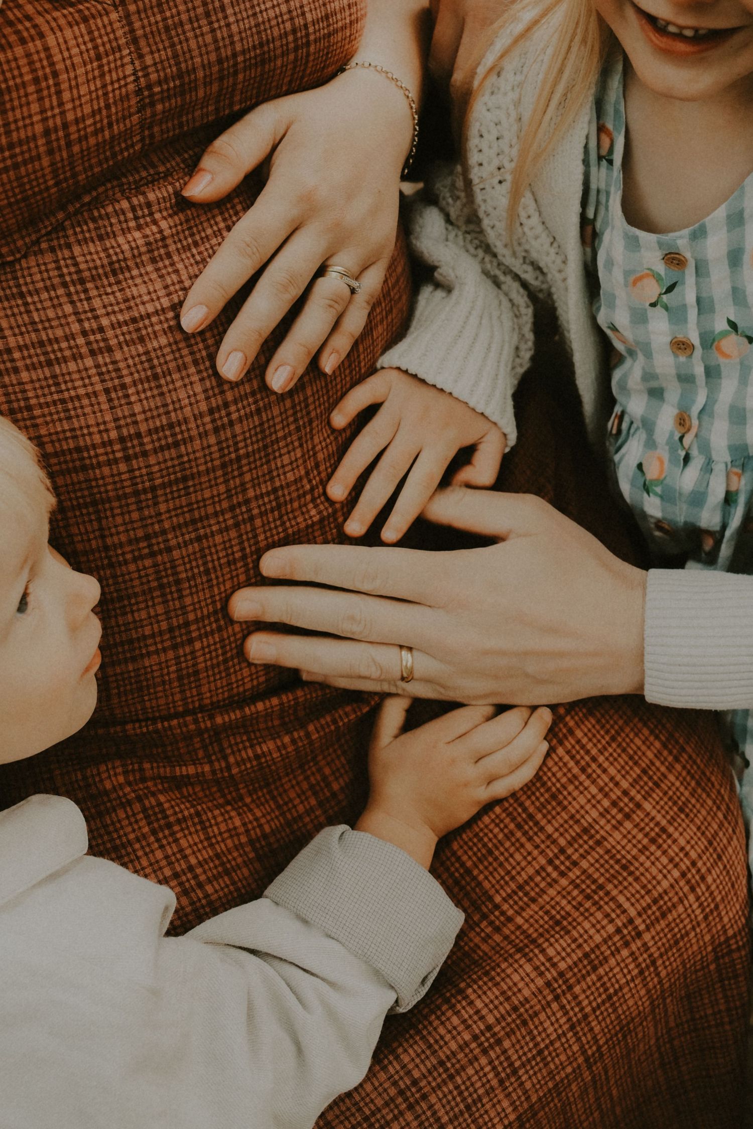 A mother and two children placing hands together on a brown plaid blanket, wearing rings and casual clothing.