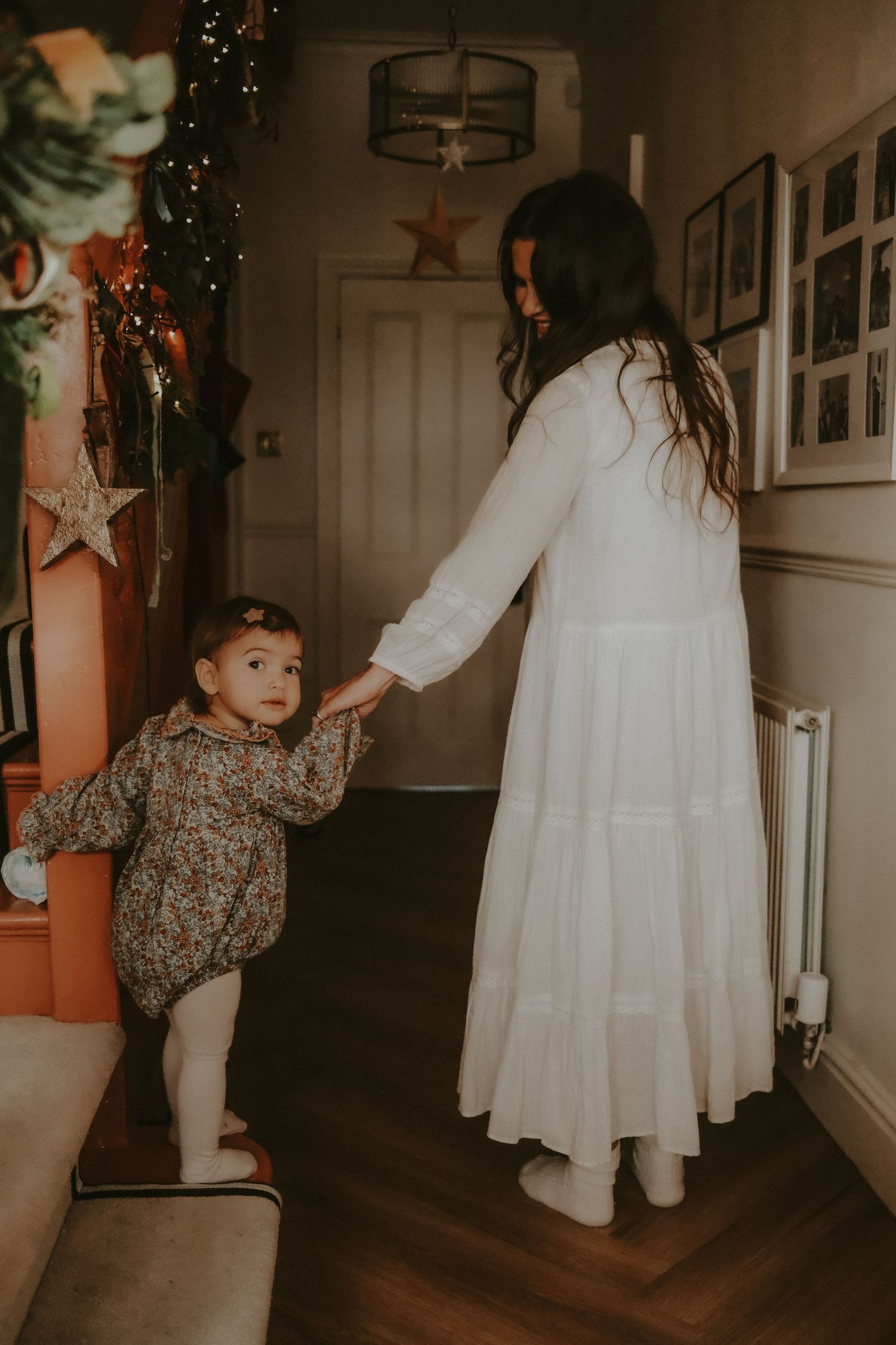Mother in white dress holding hands with baby daughter in floral outfit in Christmas decorated hallway.