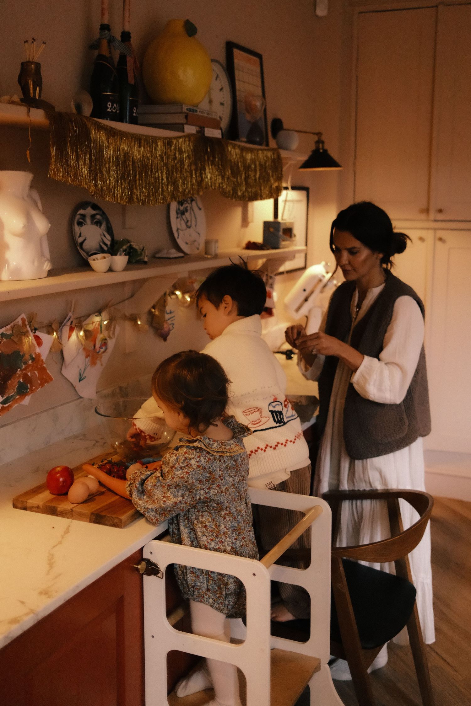 Mother and two young children cooking together in a warmly lit festive decorated kitchen.