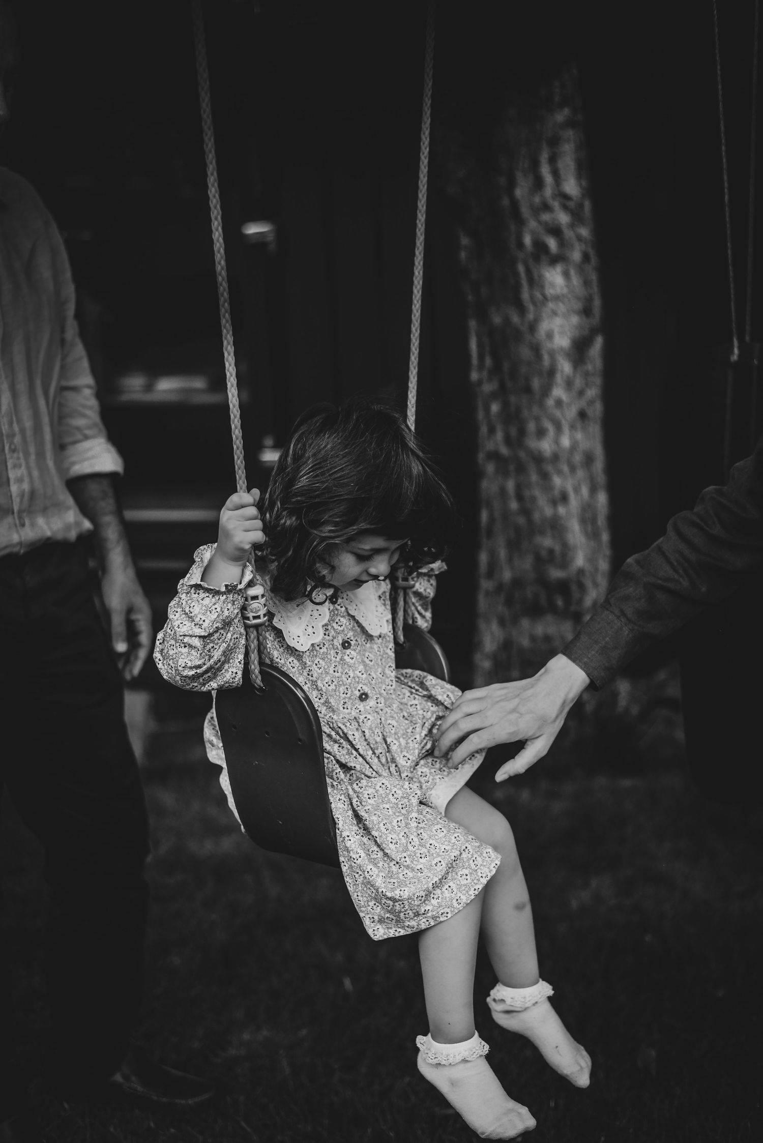 Black and white photo of a young girl on a swing being pushed by two adults outdoors.