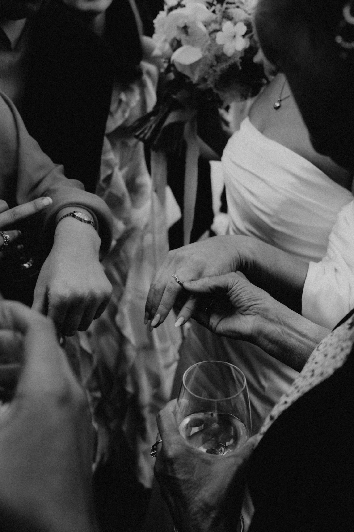 Black and white wedding photo of bride holding bouquet and champagne glass while guests admire her ring.