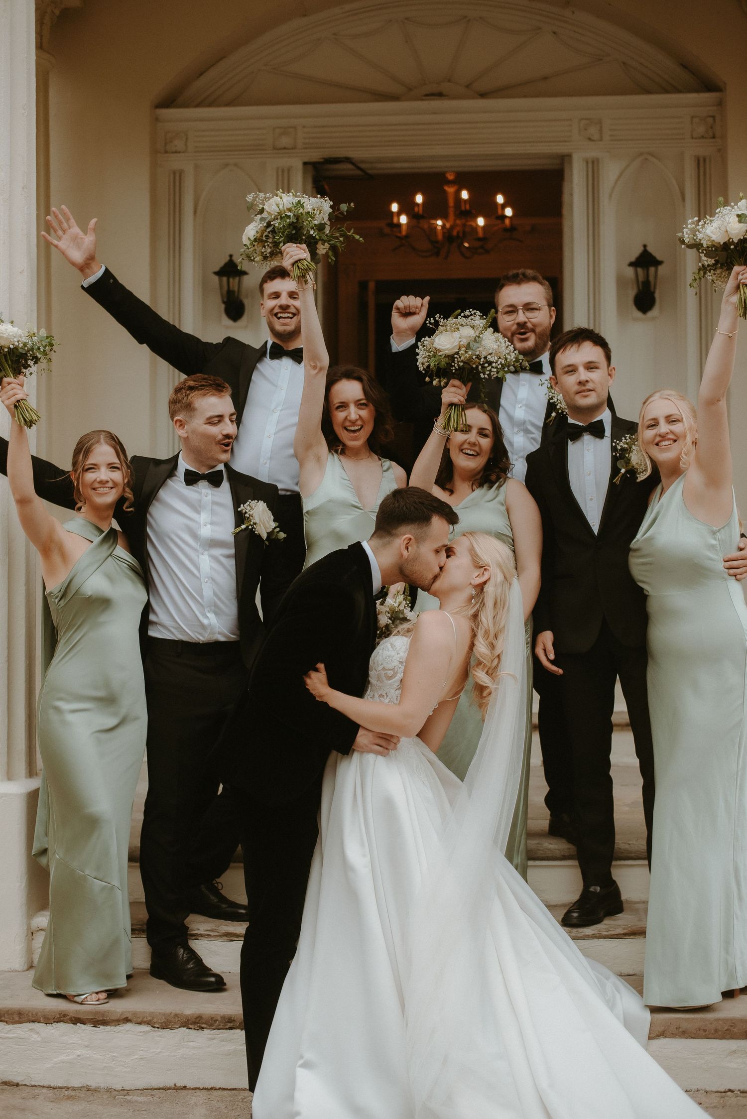 Bride and groom share a kiss surrounded by cheering wedding party in formal attire on elegant venue steps.