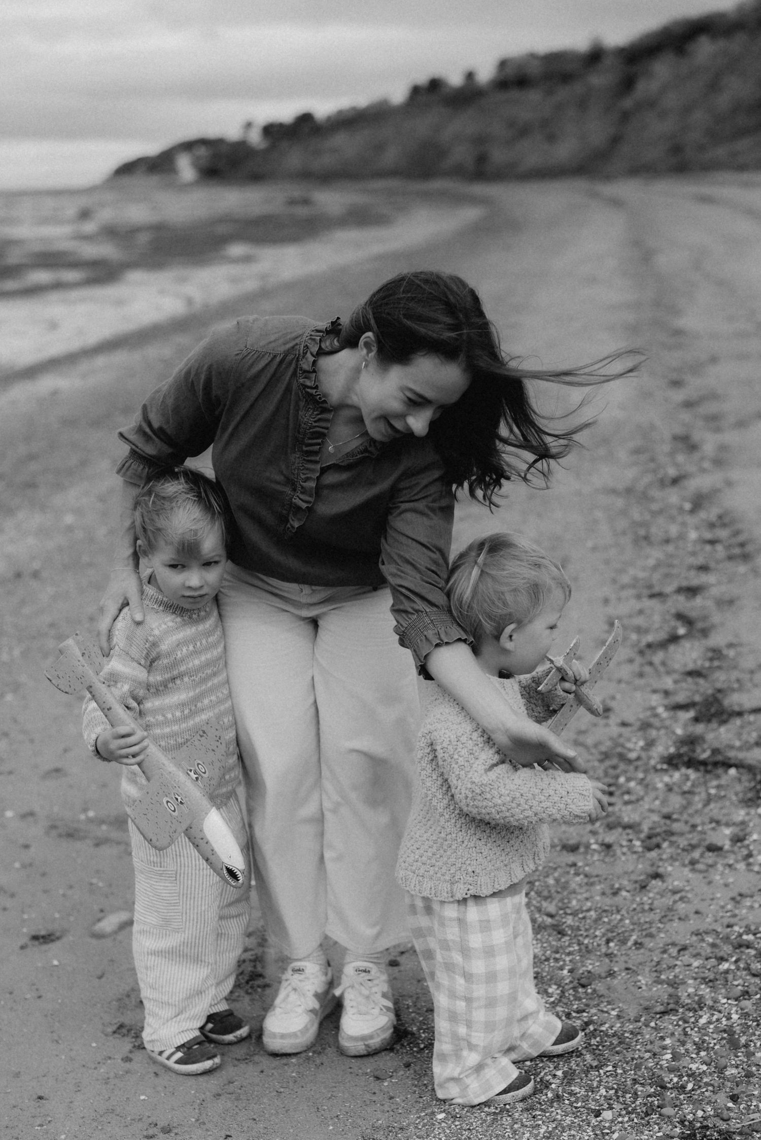 Black and white photo of a mother smiling at two young children on a beach, all holding sticks.