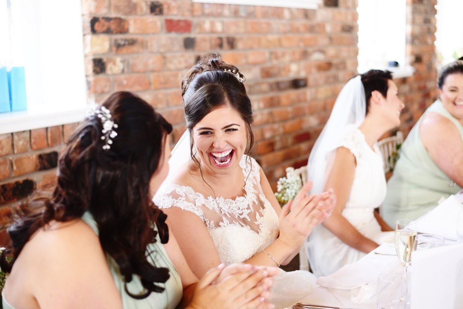 Bride in white lace gown laughing joyfully with bridesmaids in mint dresses at a rustic brick wall venue.