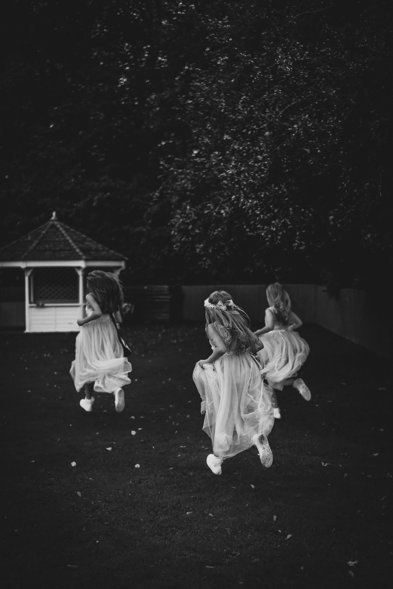 Three young girls in white dresses run joyfully across a garden lawn in a black and white photo.