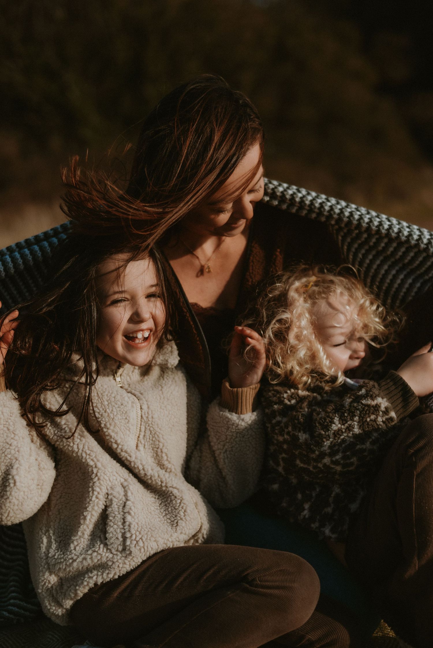 Warm outdoor portrait of a mother smiling with two young children wrapped in a striped blanket.