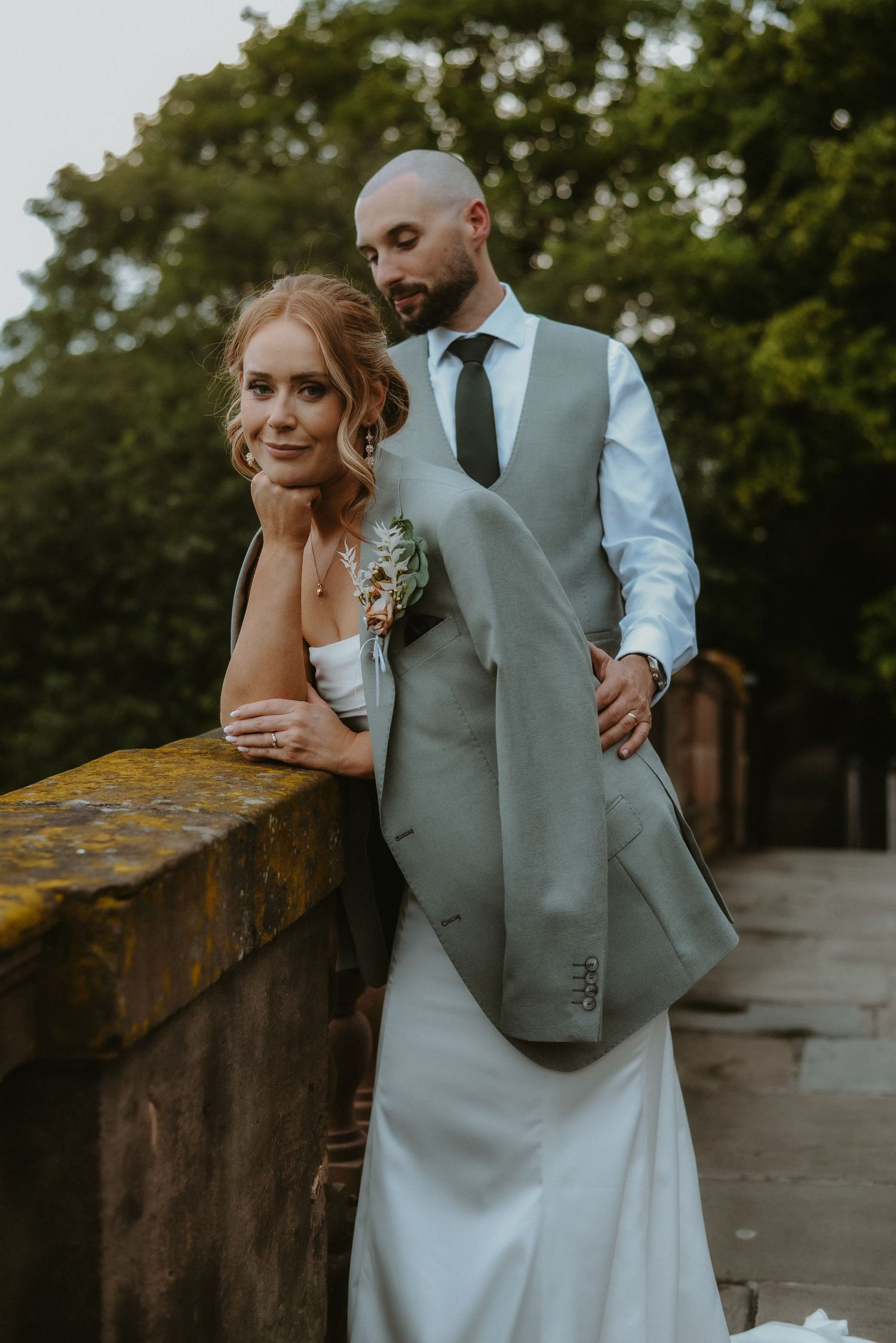 Bride in white gown and groom in grey suit pose romantically on a stone bridge surrounded by lush greenery.