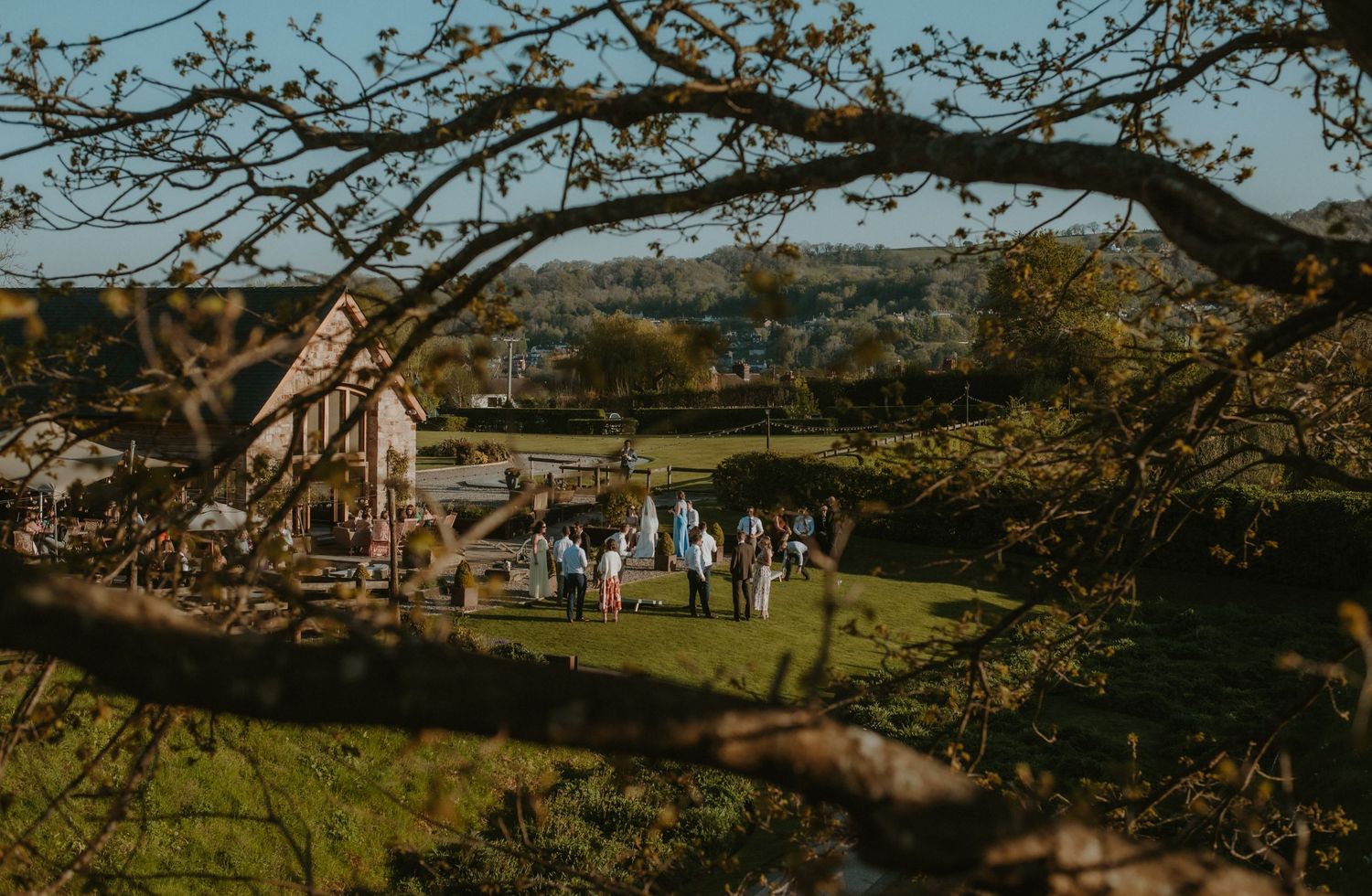 Guests gather on lush green lawn beside rustic barn with scenic countryside views framed by spring tree branches.