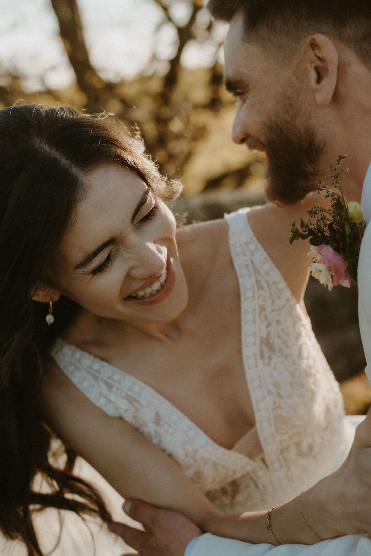 A smiling bride in a lace wedding dress embracing her bearded groom outdoors.