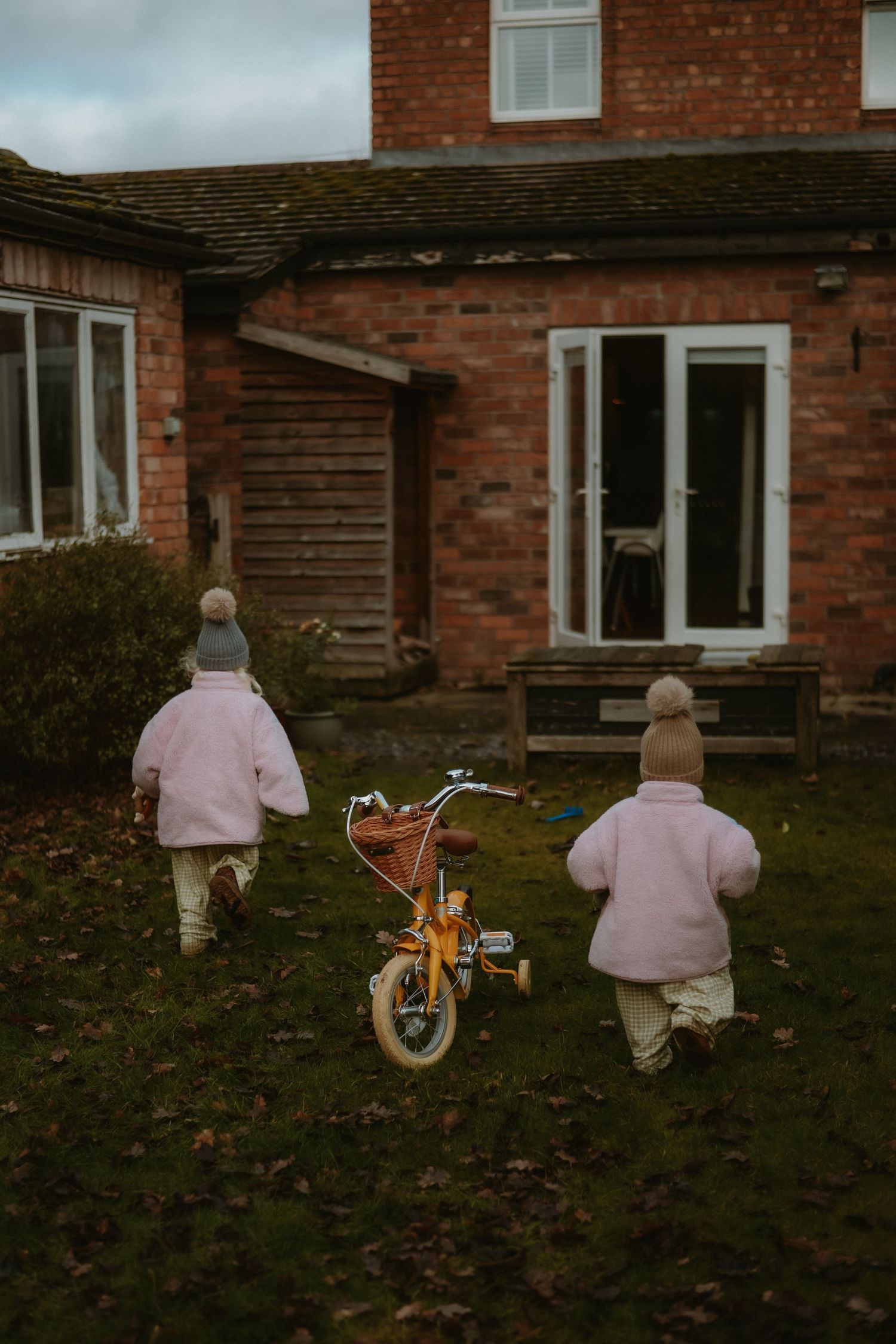 Two young children in pink coats walking toward a red brick house with a yellow bicycle.