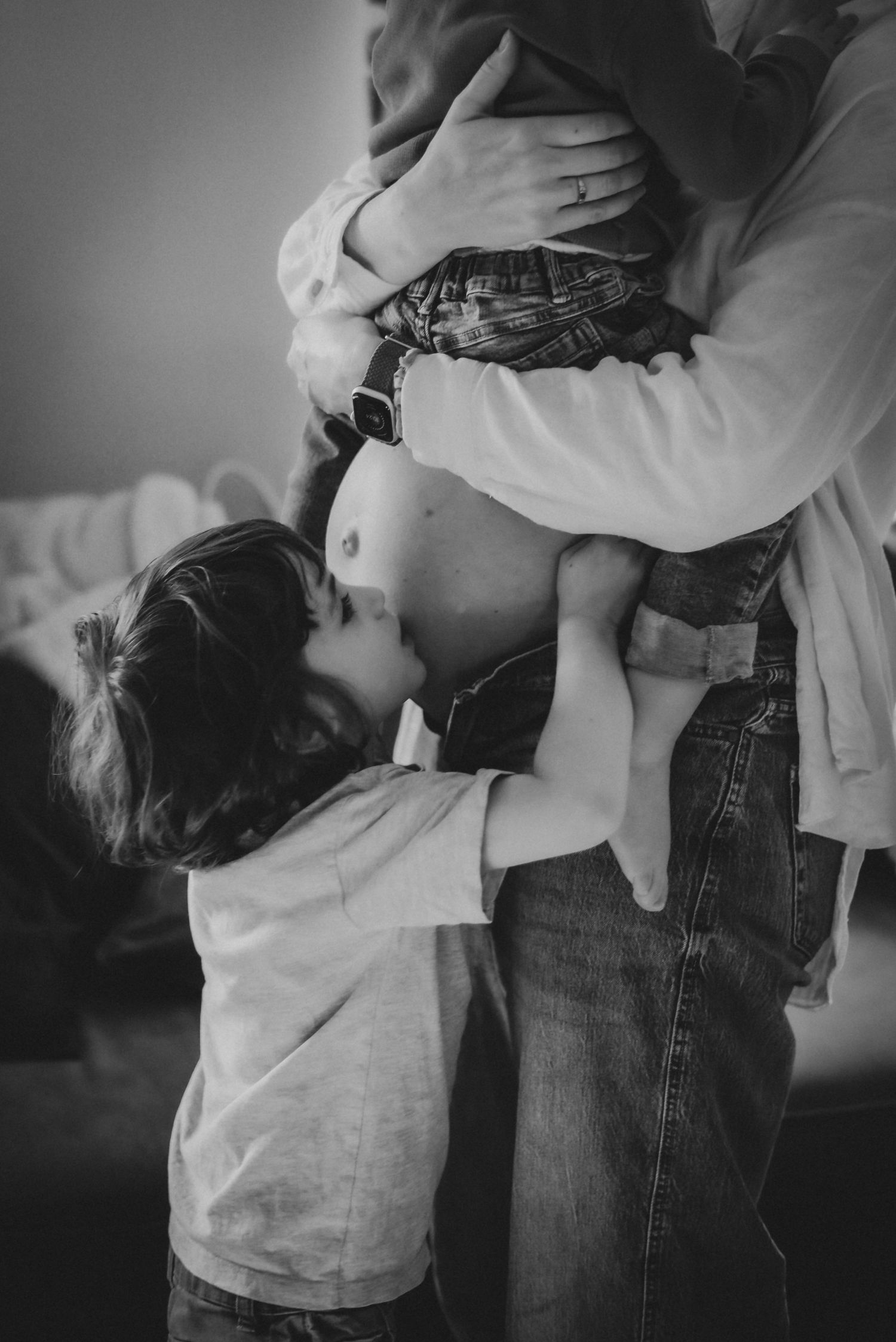 Black and white photo of a young child kissing a pregnant mothers belly lovingly.