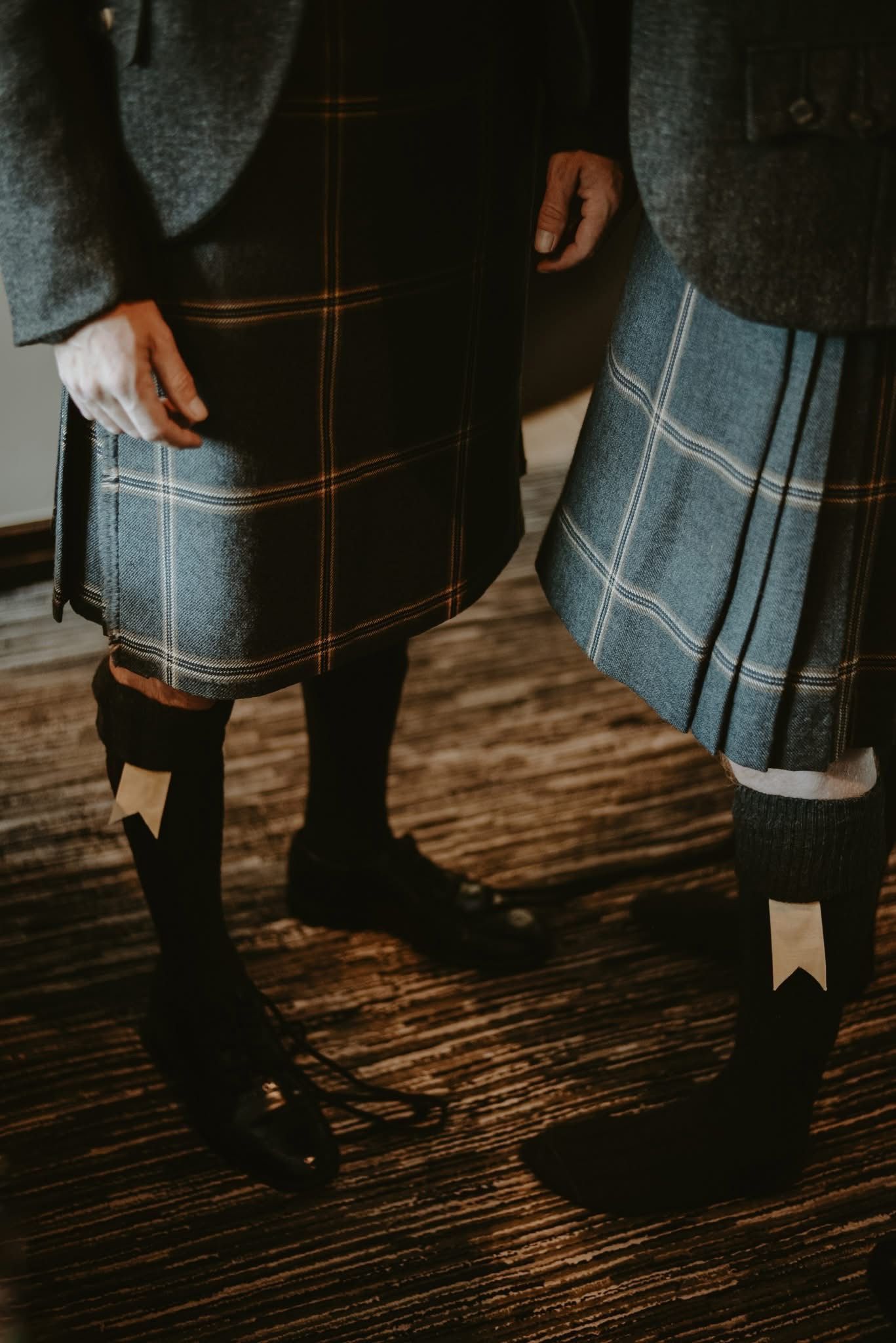 Two men wearing traditional Scottish tartan kilts and black shoes indoors.