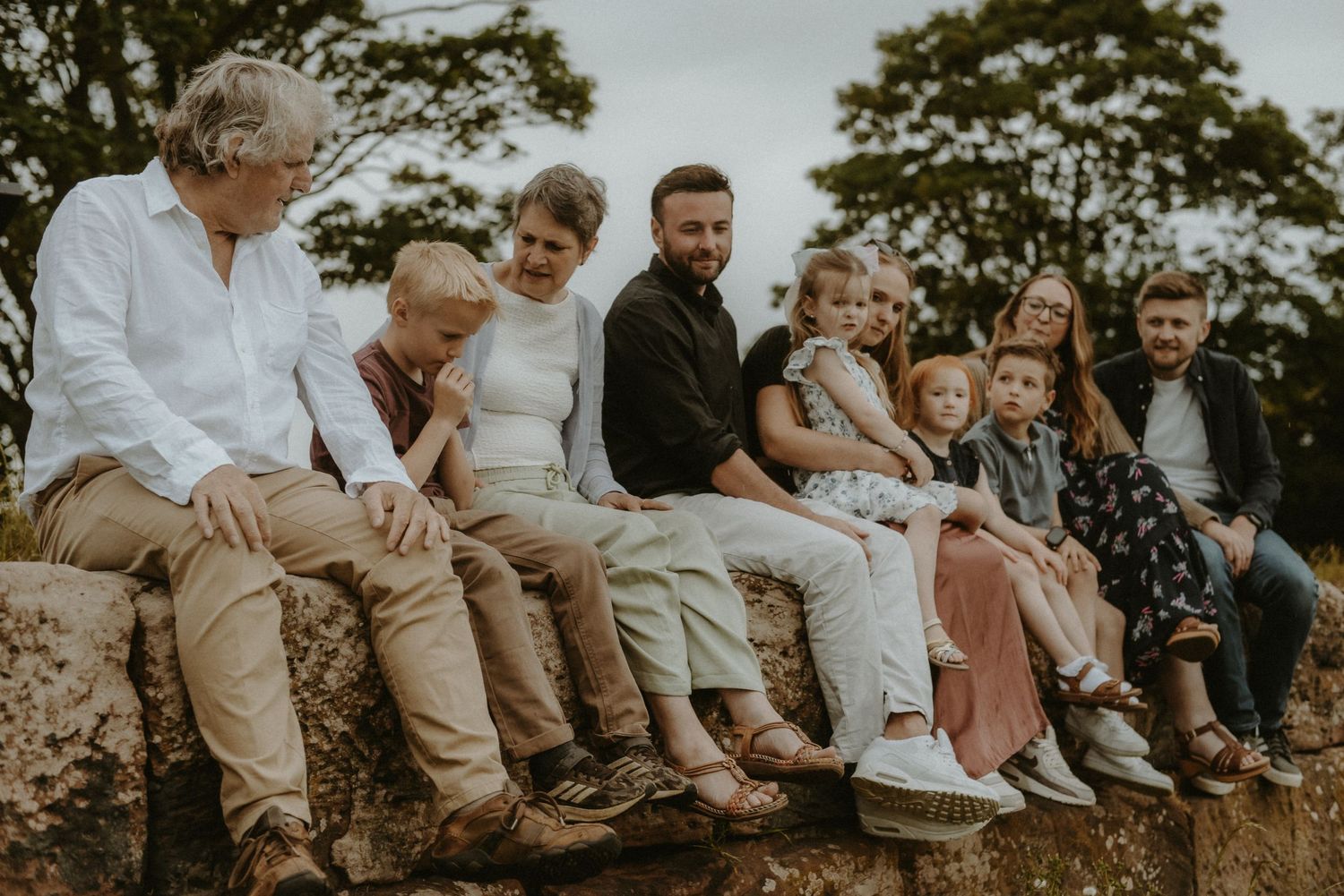 Large multigenerational family sitting together on a stone wall outdoors, smiling and bonding in a scenic natural setting.