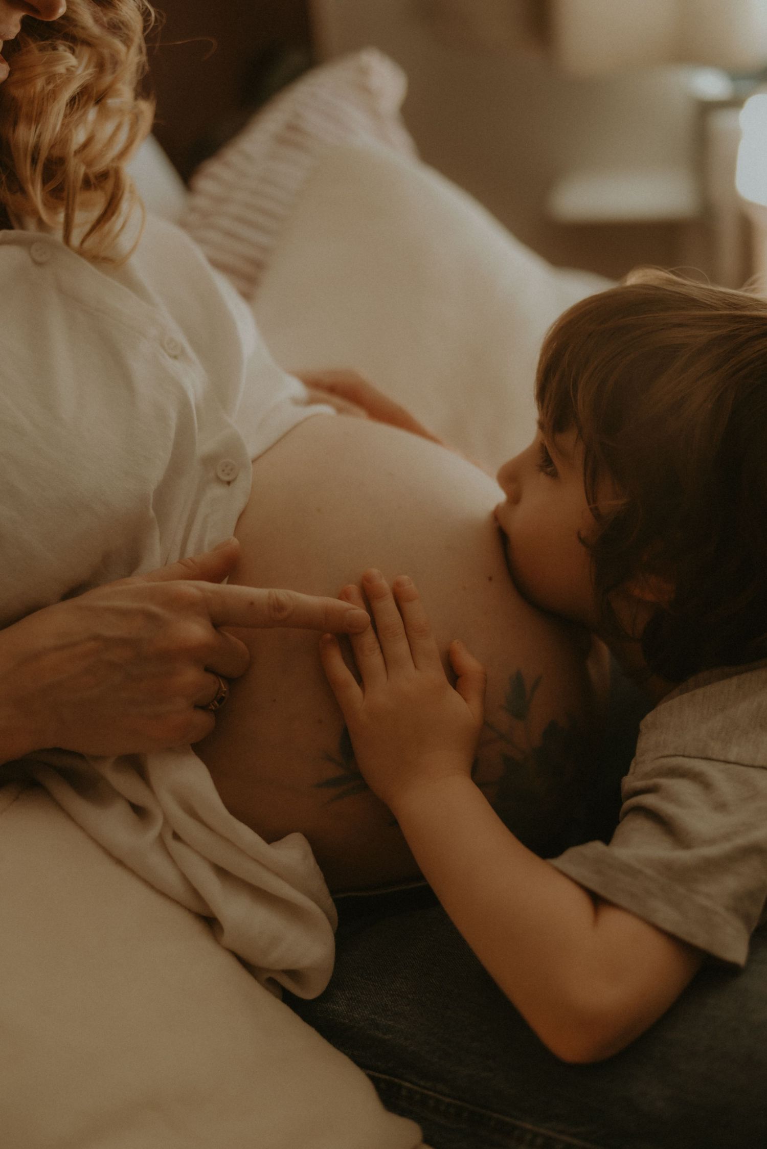 A young child gently touches a pregnant woman's bare belly while resting on a cozy bed with white pillows.