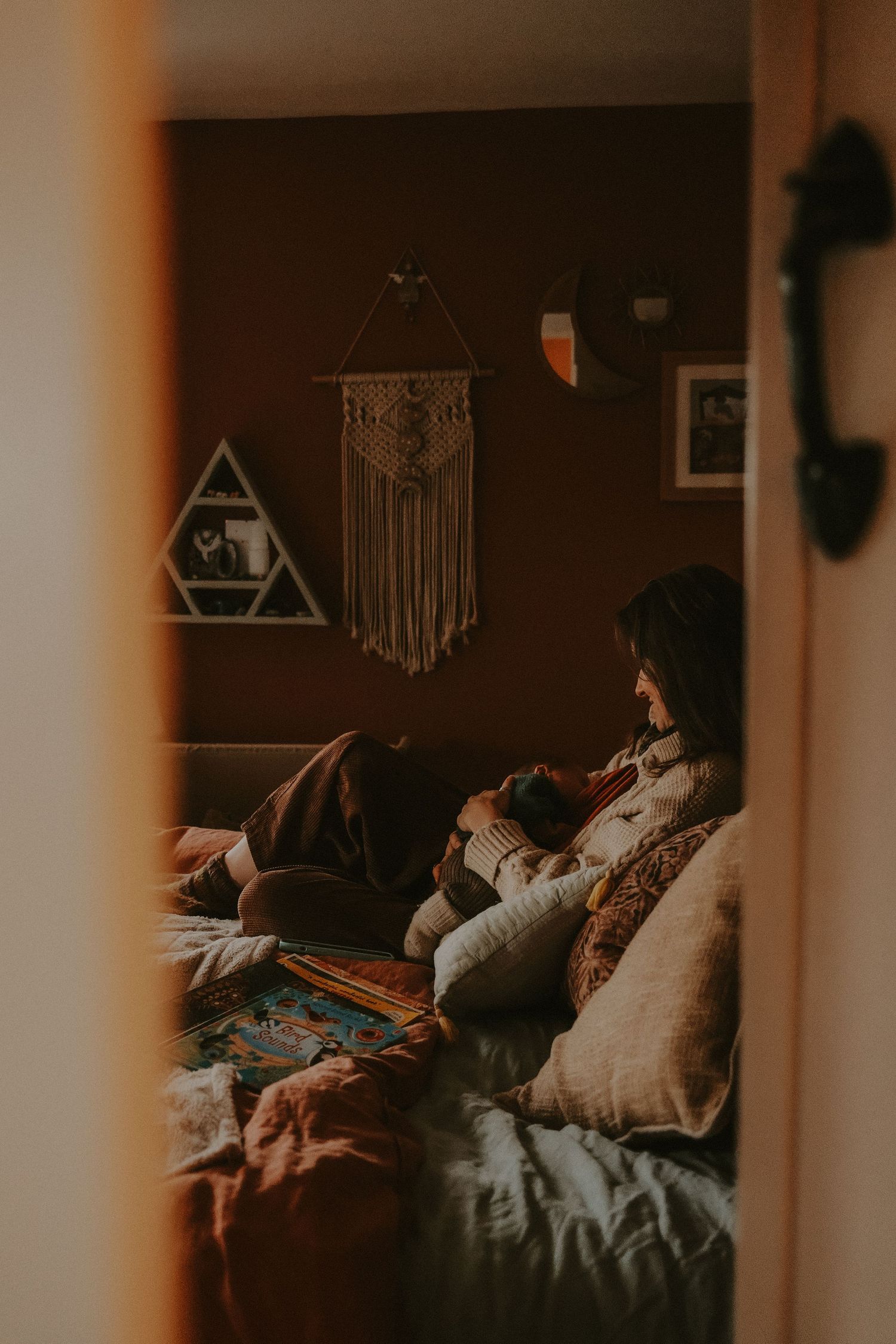A woman relaxes on a boho-styled bed holding a baby, with macrame wall decor in a warmly lit bedroom.