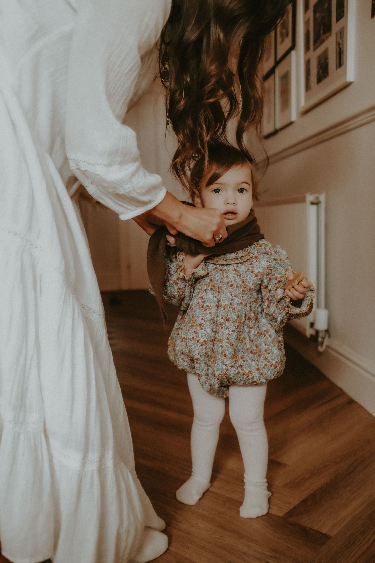 A curious baby girl in a floral romper standing with support from her mother indoors.