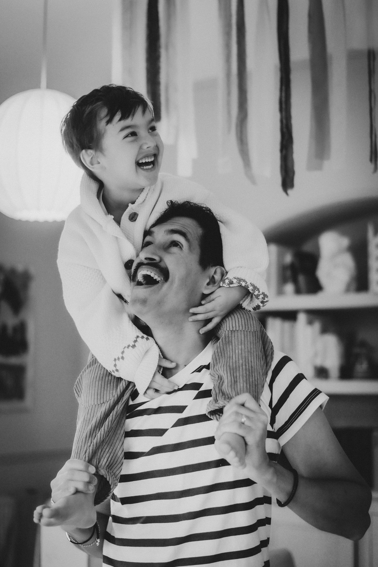 Black and white photo of a laughing child sitting on a man's shoulders indoors.