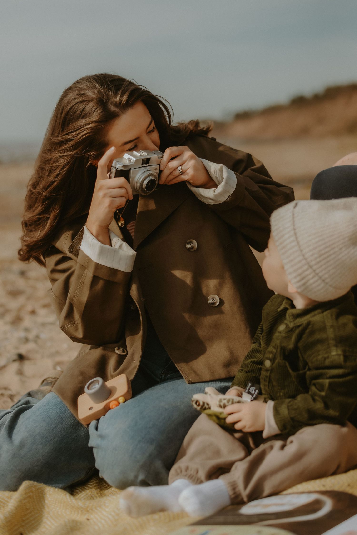 Young mother photographing toddler with vintage camera during beach picnic outing.