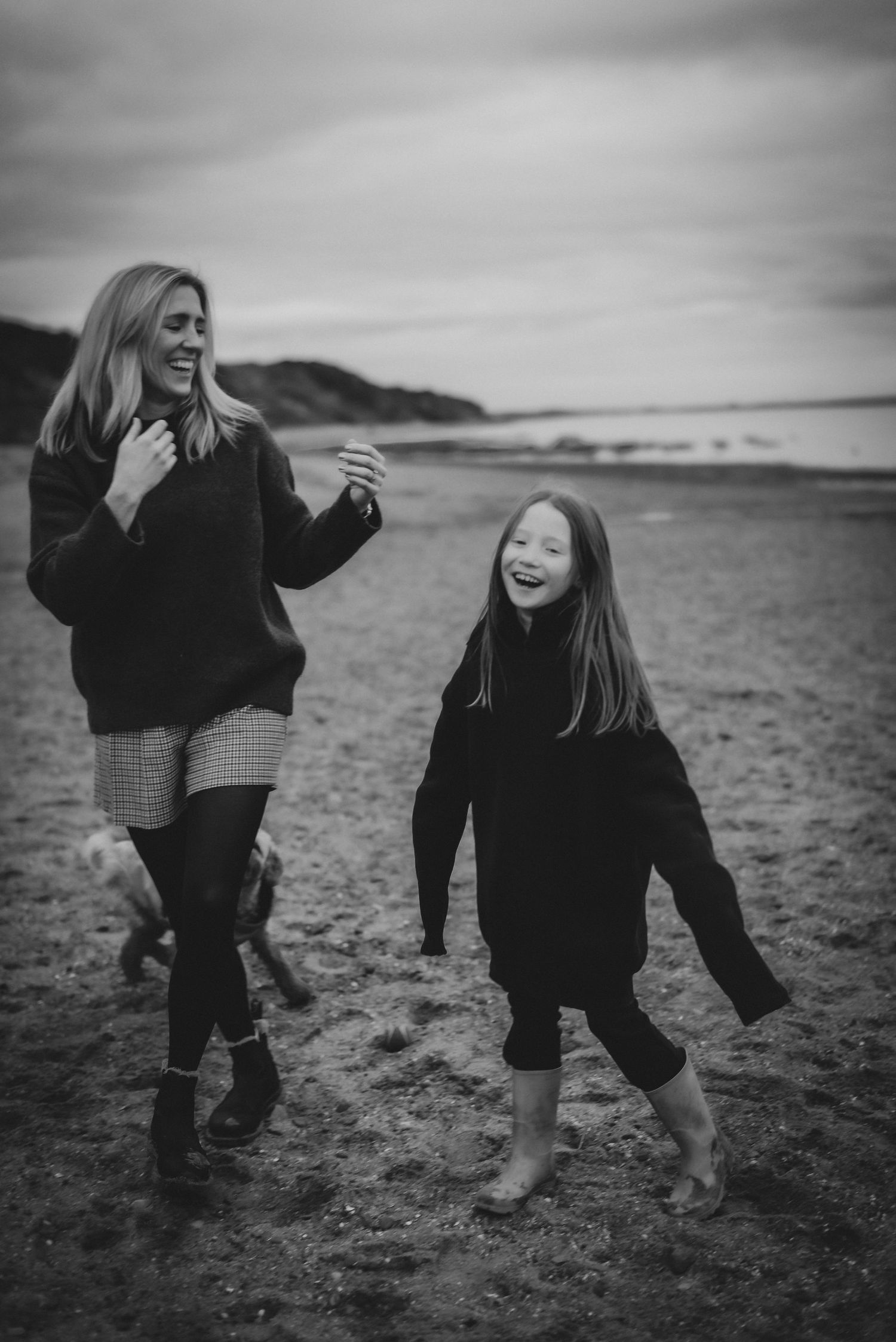 Black and white photo of laughing mother and daughter walking barefoot on beach.