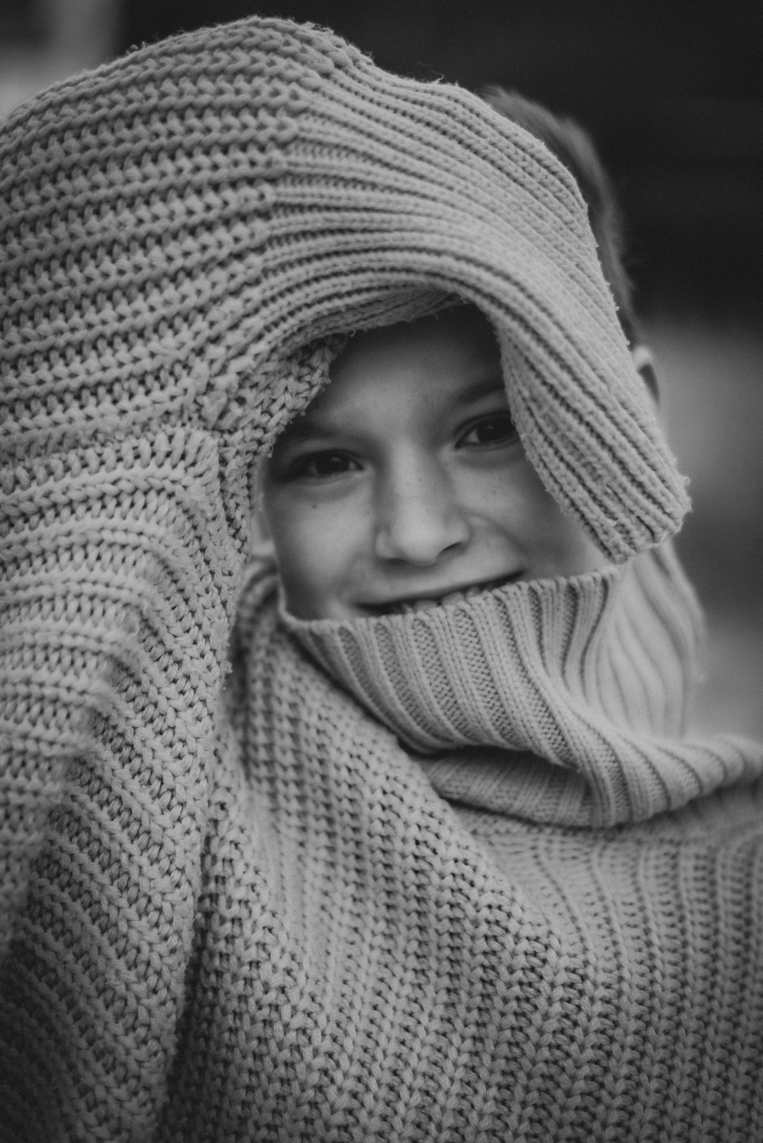 Black and white portrait of a smiling child playfully wrapped in a large chunky knit sweater.
