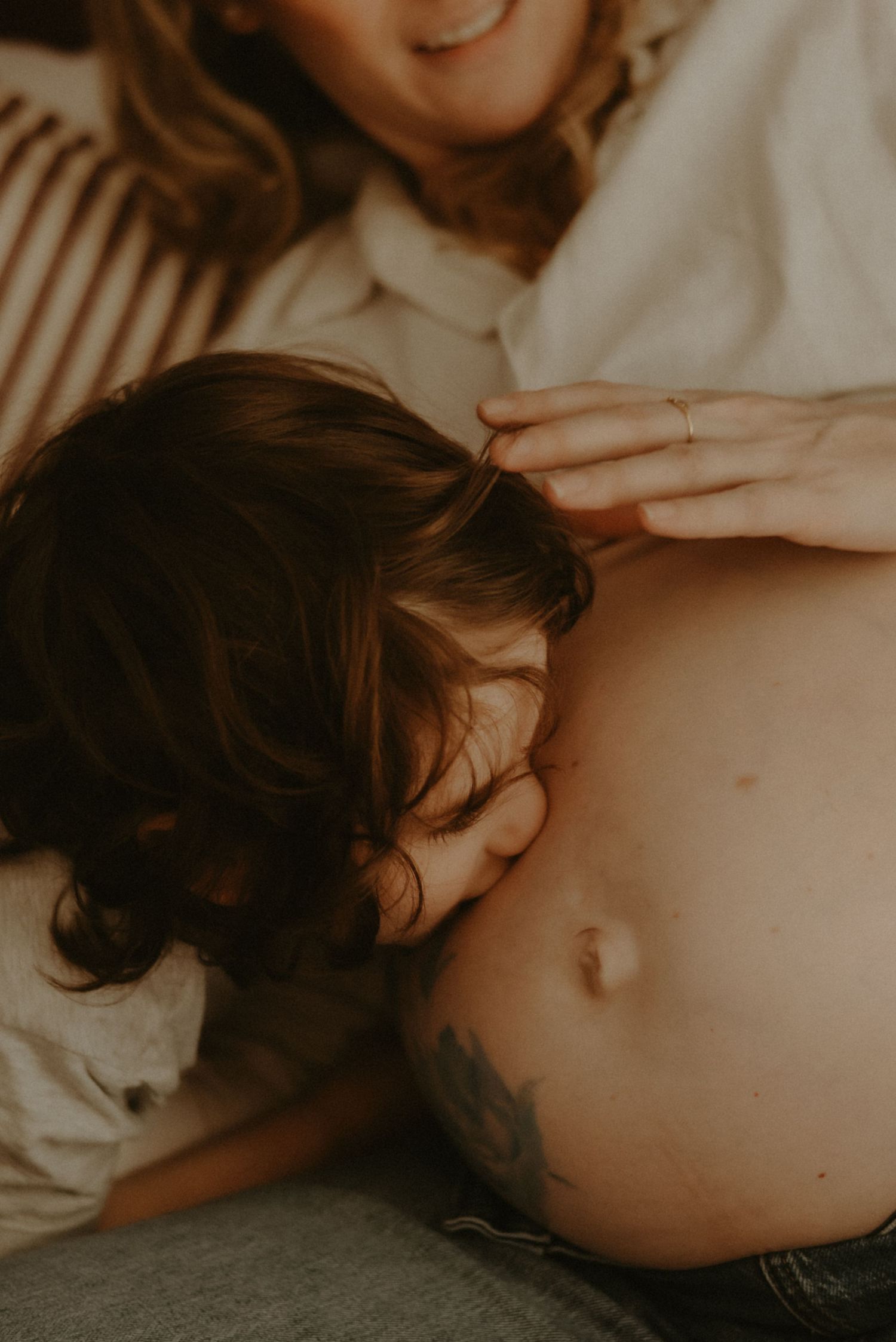 A child resting their head on a pregnant woman's bare belly in a warm, intimate moment.