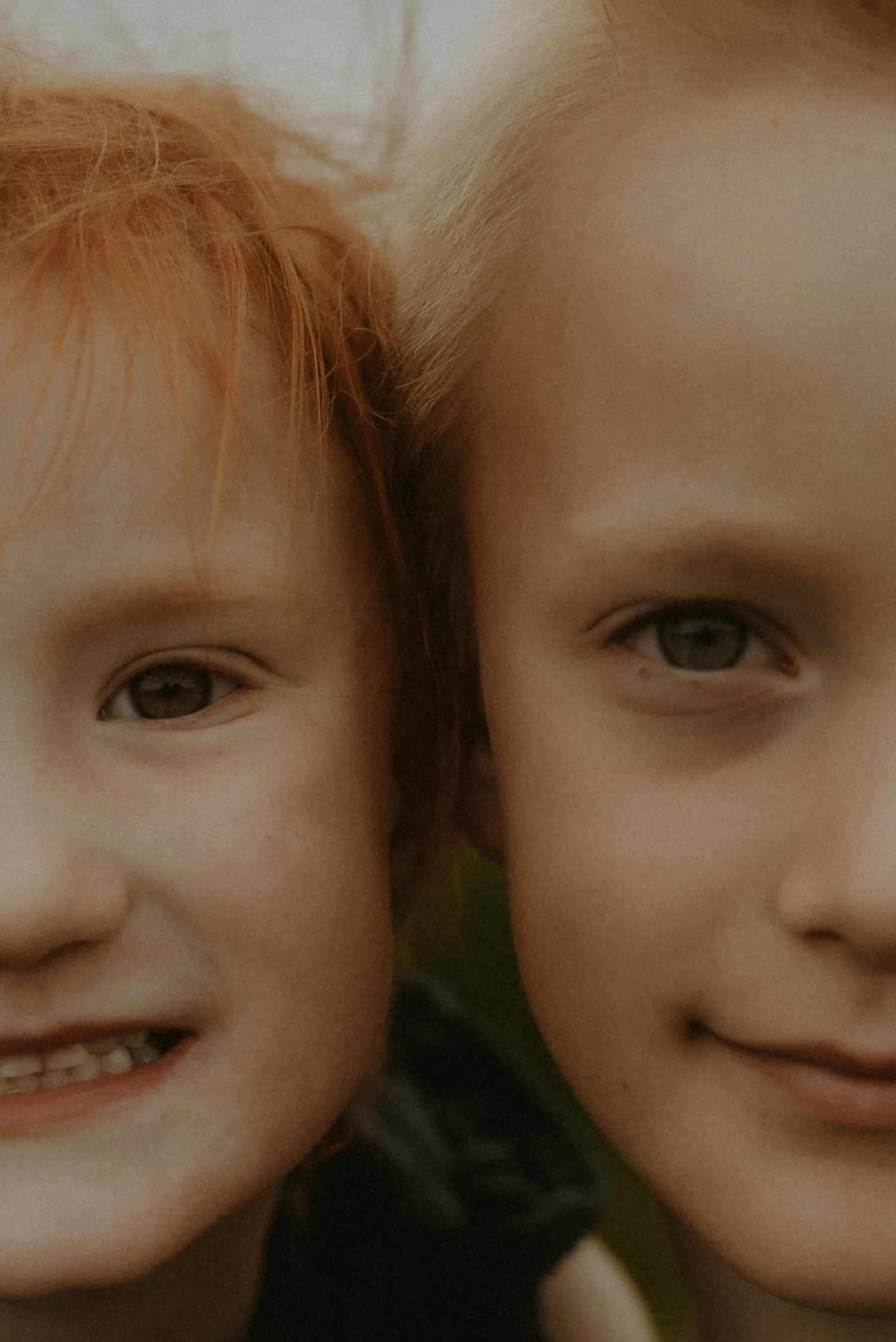 Close-up portrait of two smiling red-haired children with freckles and green eyes posing cheek to cheek outdoors.