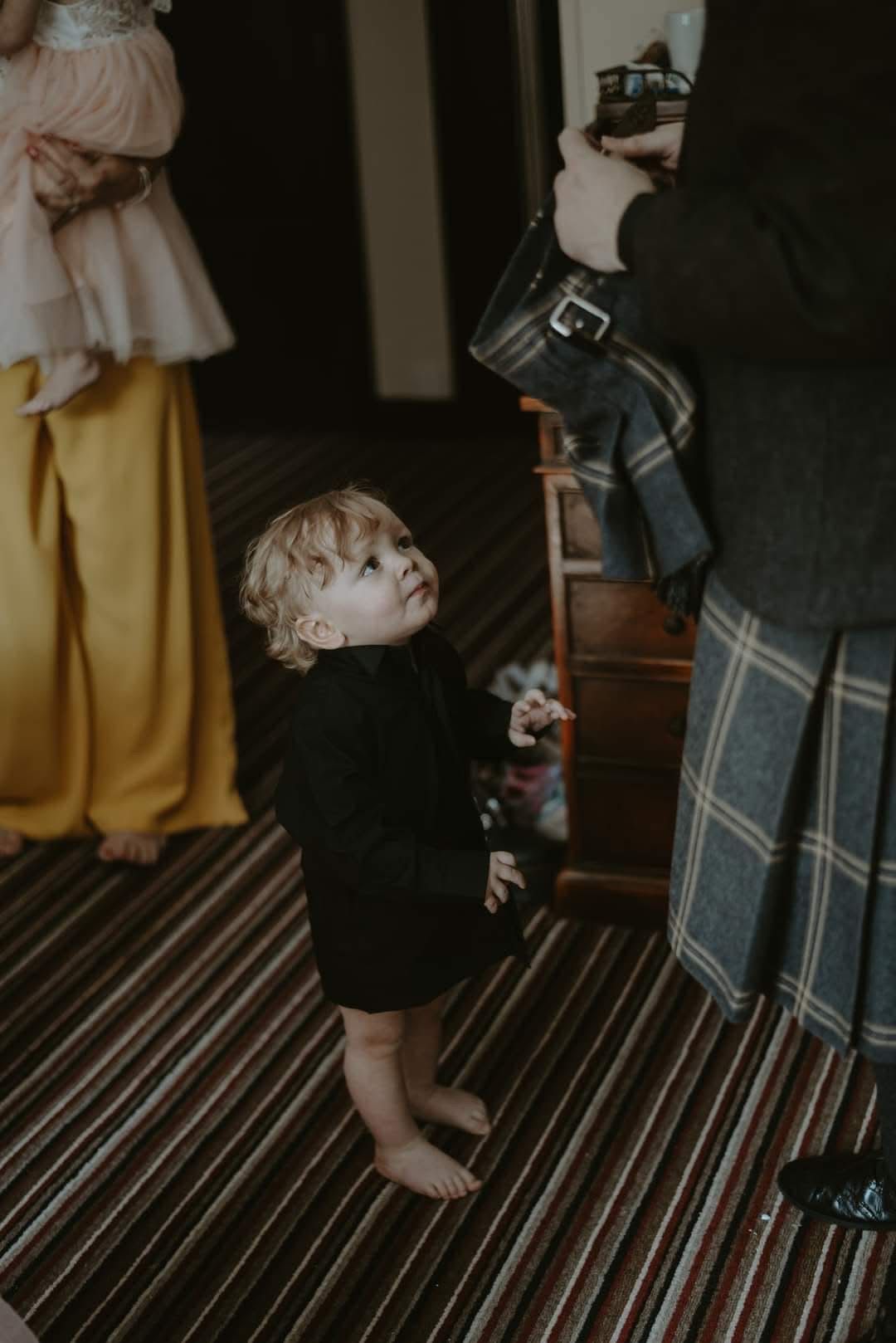A barefoot toddler in a black outfit looks up curiously at an adult in a plaid kilt during a wedding event.