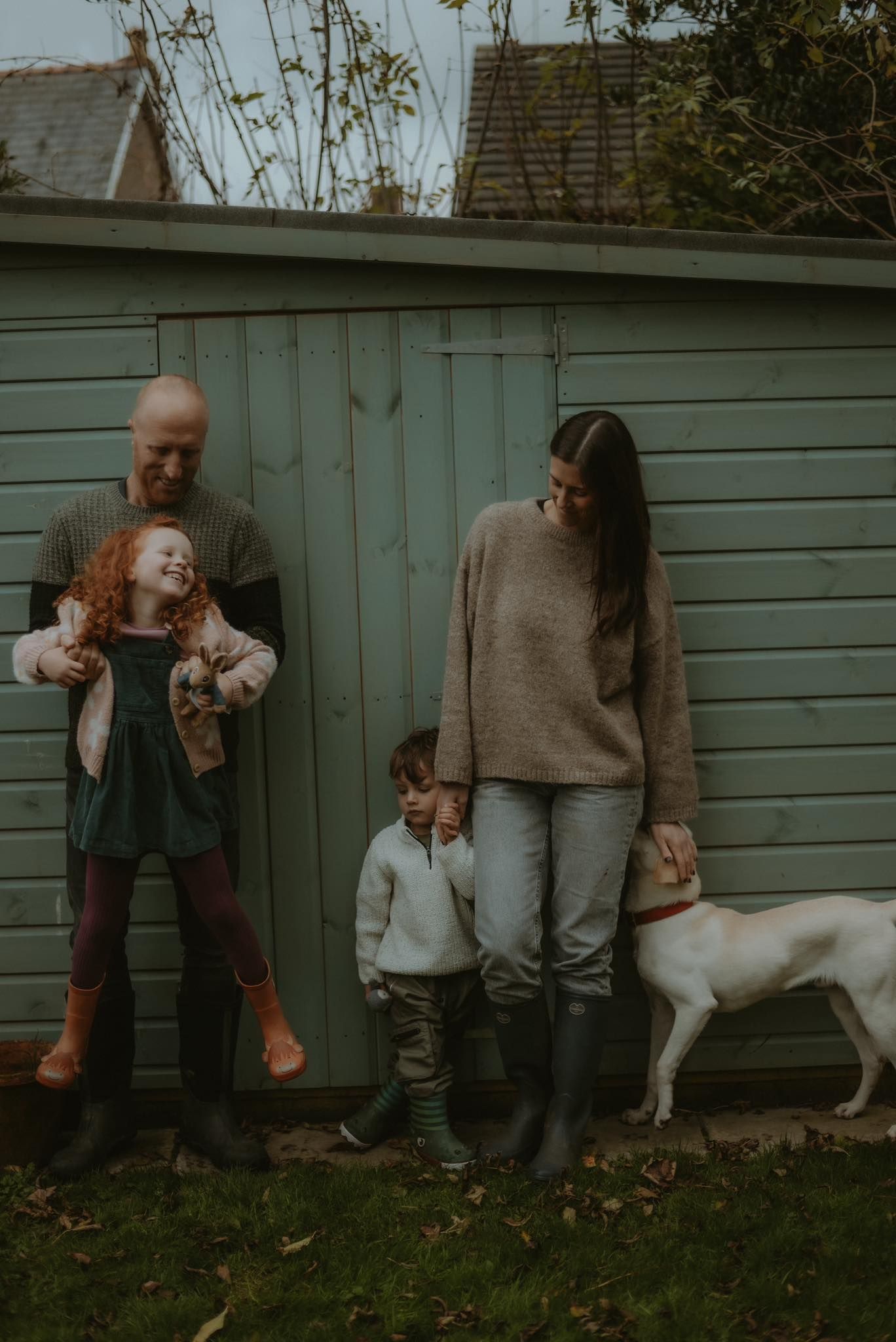 Family of four with a white dog pose casually against a sage green garden shed outdoors.