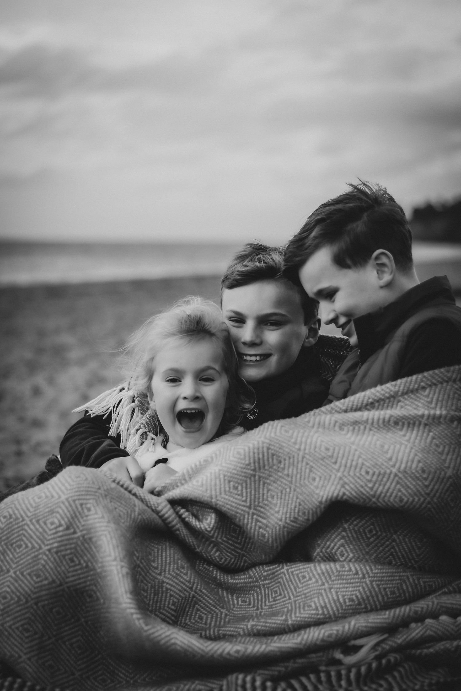 Black and white photo of three siblings laughing together wrapped in a blanket on a beach.